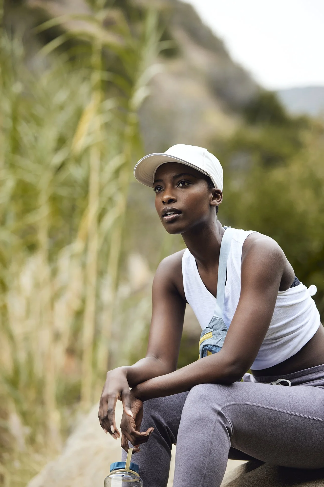 A young woman with dark skin wearing a white sports cap, sleeveless white top, and grey joggers, sits outdoors holding a water bottle, with lush foliage in the background.