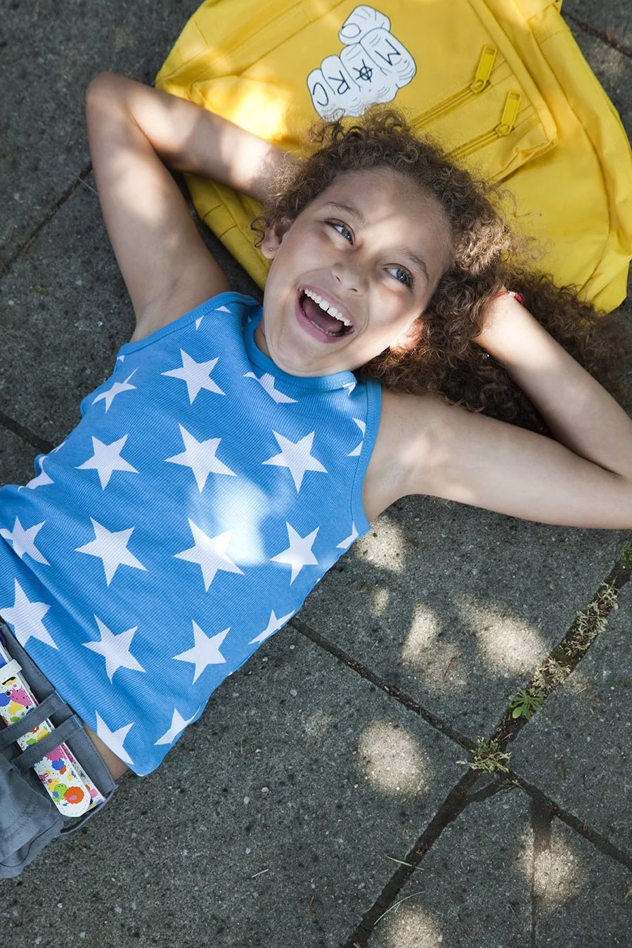 A young girl with curly hair lying on a concrete sidewalk, smiling and looking up, wearing a blue sleeveless shirt with white stars, with a yellow bag nearby.