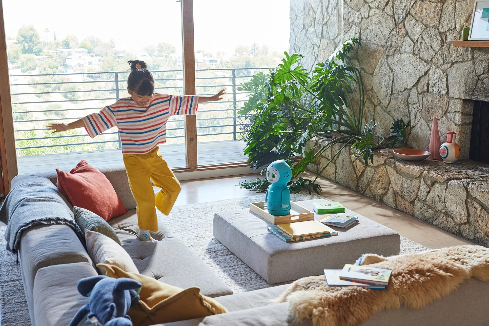 A young girl jumping on a beige sofa in a living room with a large window showing a scenic outdoor view. The room has a stone fireplace, green plants, decorative vases, and a coffee table with books and a blue robot toy.