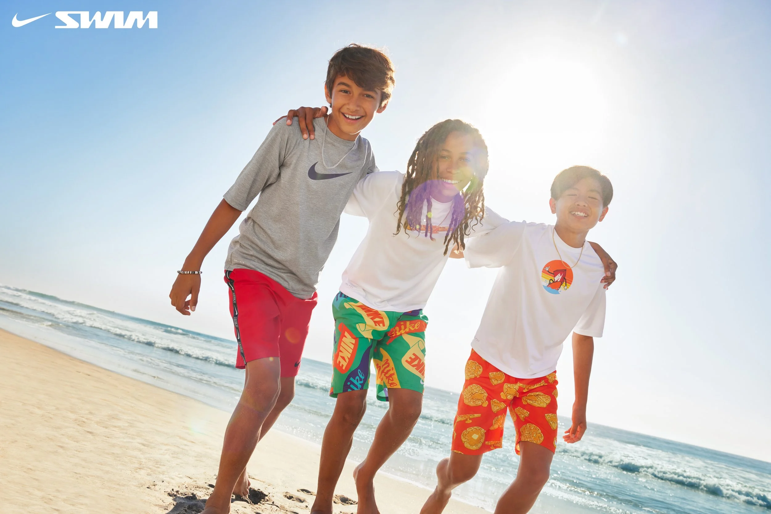 Three children smiling and playing on the beach near the water, with the sun shining brightly in the sky.