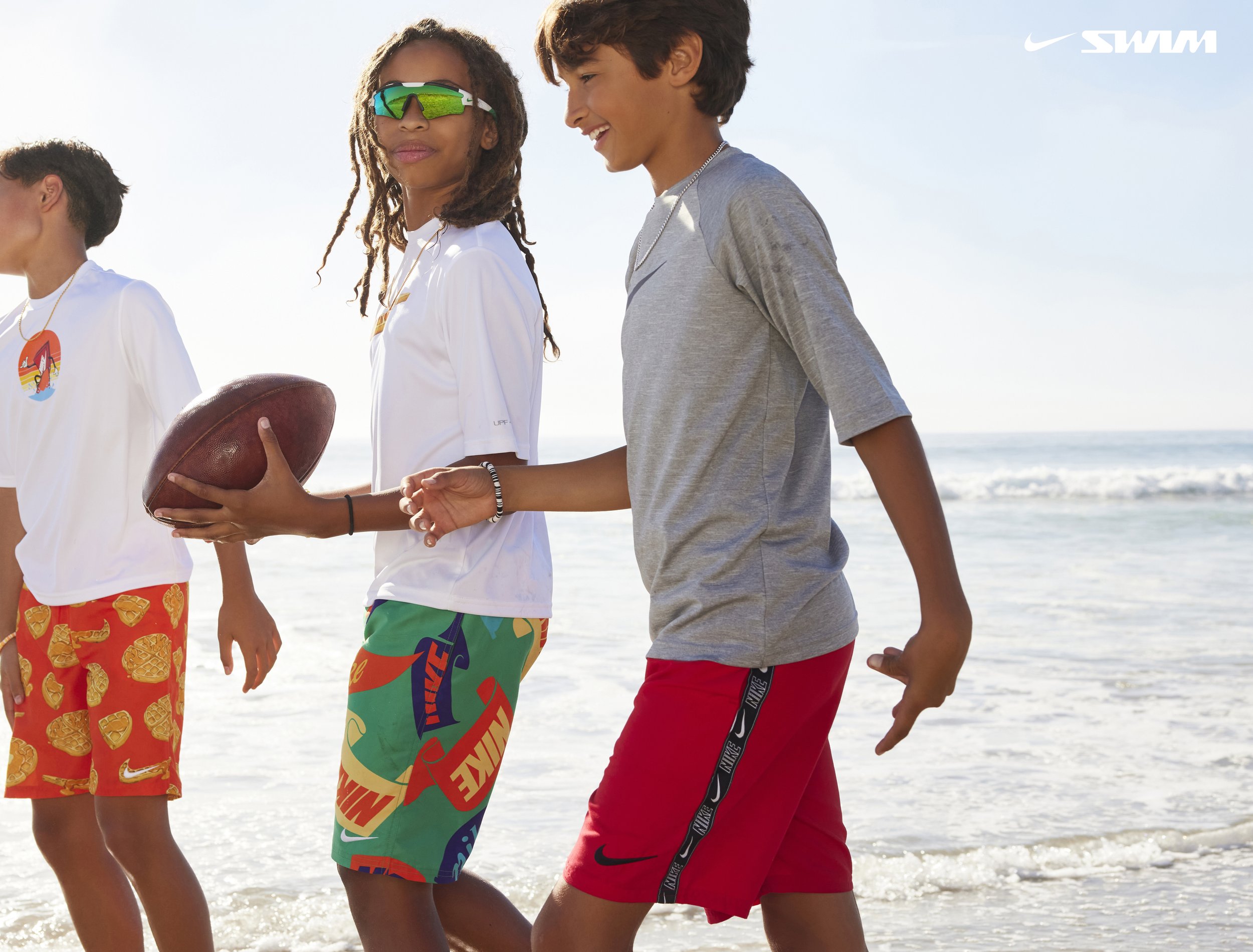 Four teenagers on the beach, one holding a football, enjoying a sunny day near the ocean.
