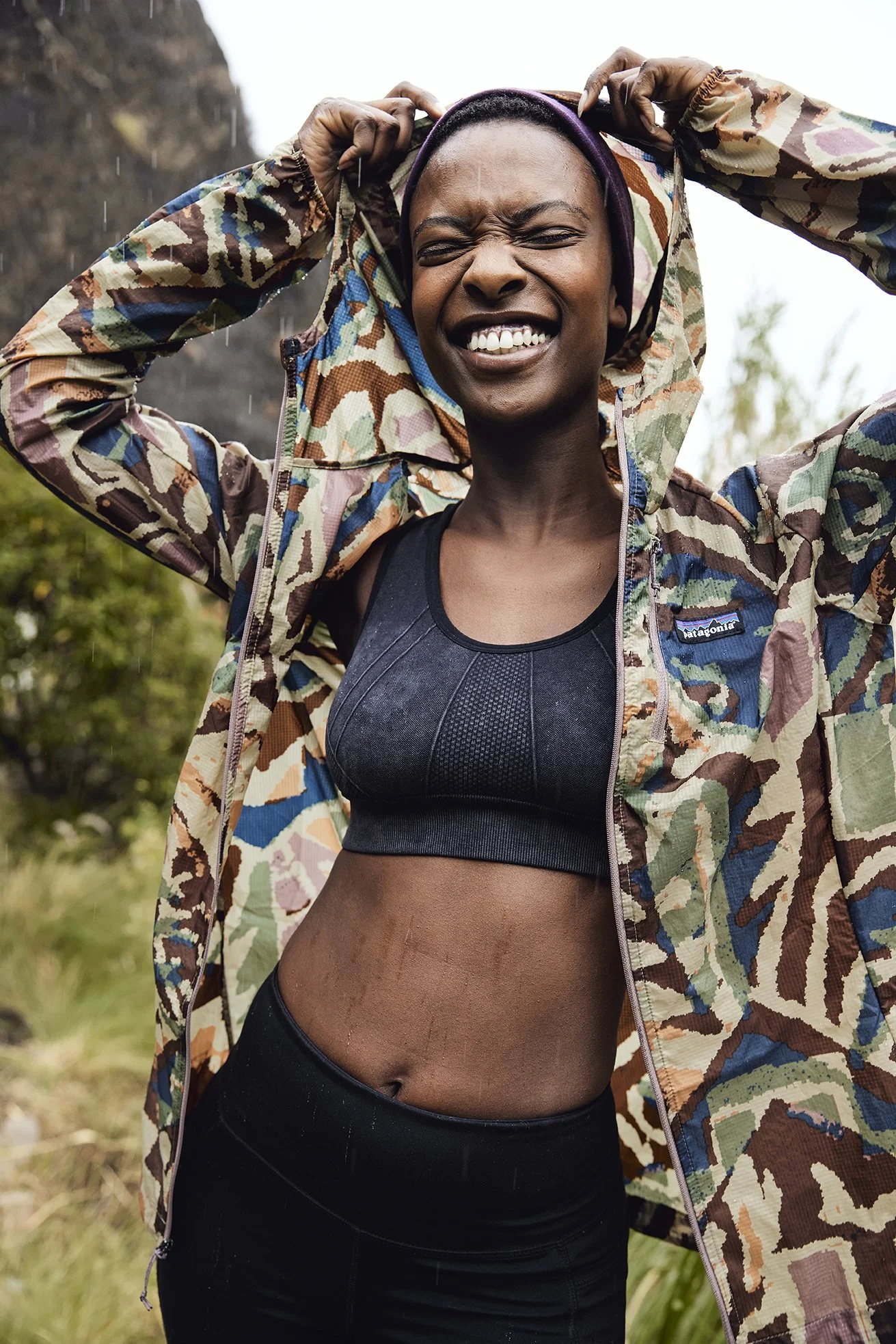 A woman outdoors in rainy weather wearing a camouflage jacket and black sports bra, smiling with eyes closed and hands adjusting her hood.