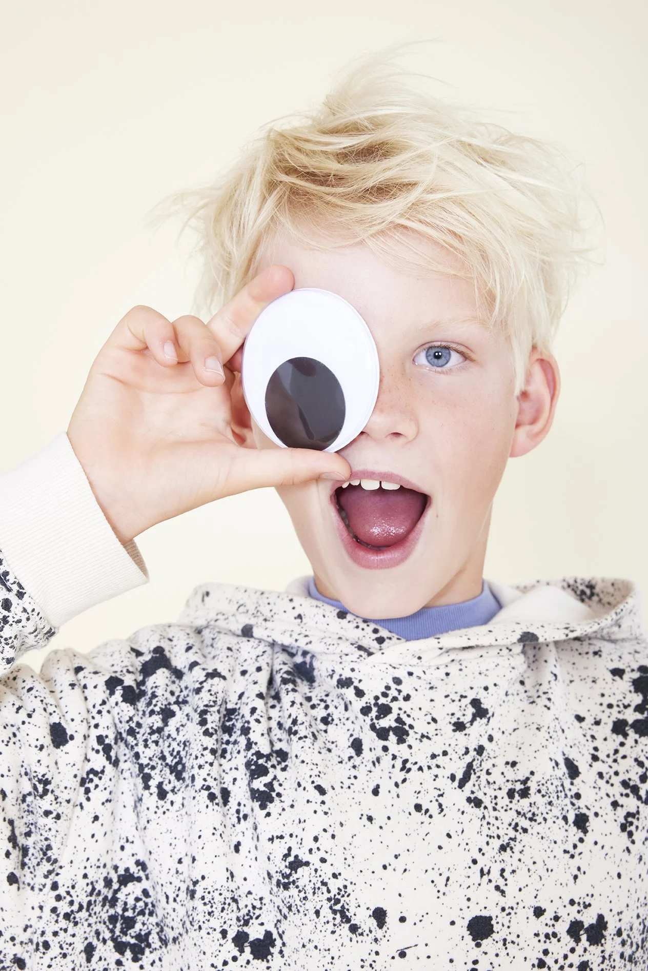 A young boy with blond hair and blue eyes holding a paper eyepatch to his right eye, making a playful expression with his mouth open.