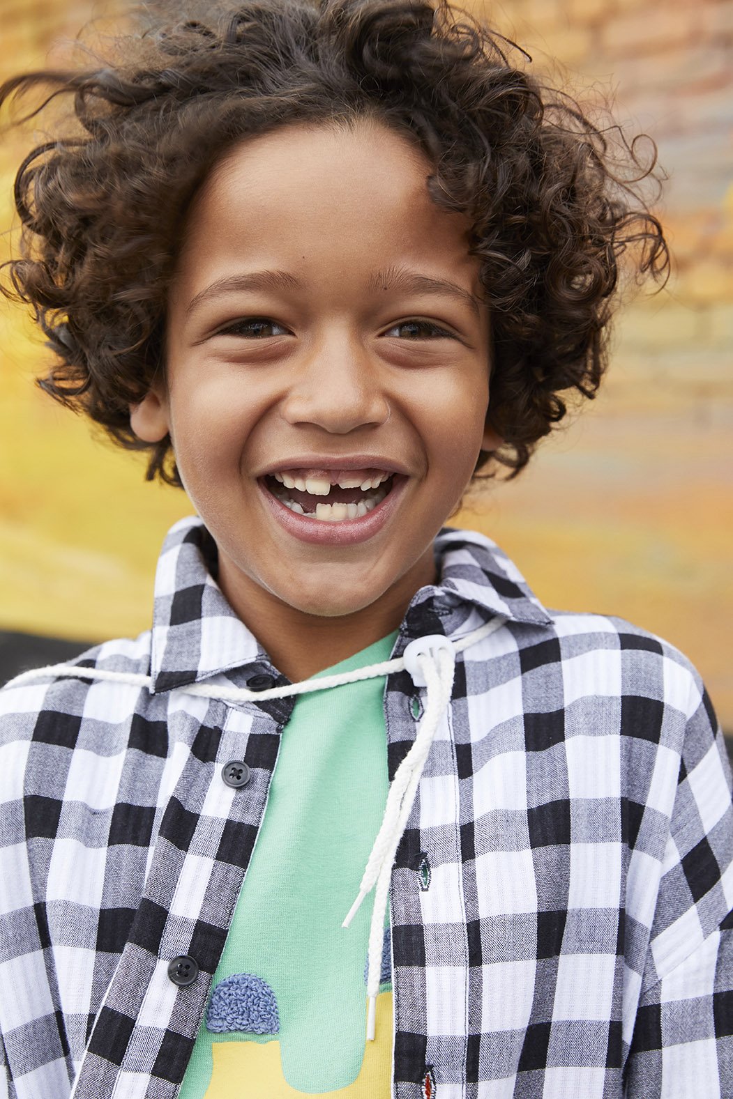 A young boy with curly hair smiling outdoors with an autumn-colored background.