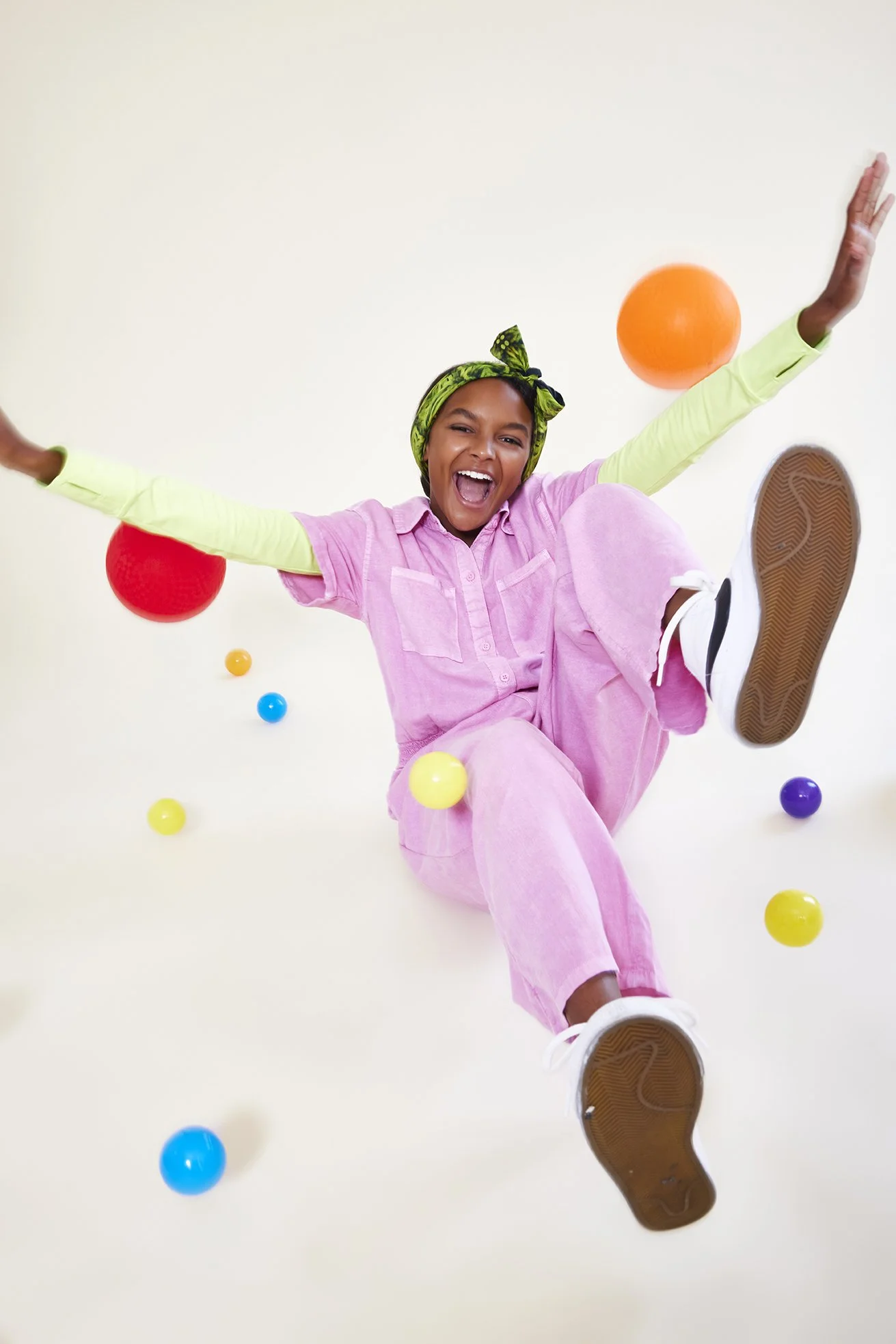 Young woman in pink outfit with a green headscarf joyfully playing with colorful balls against a plain background.