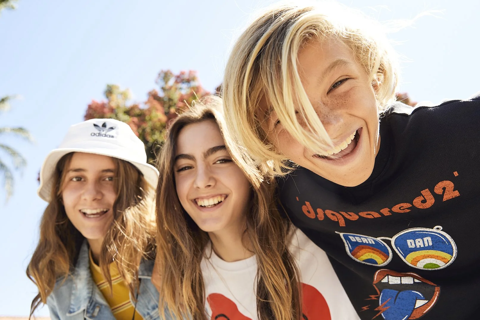 Three young friends smiling and taking a close-up selfie outdoors on a sunny day with blue sky and palm trees in the background.