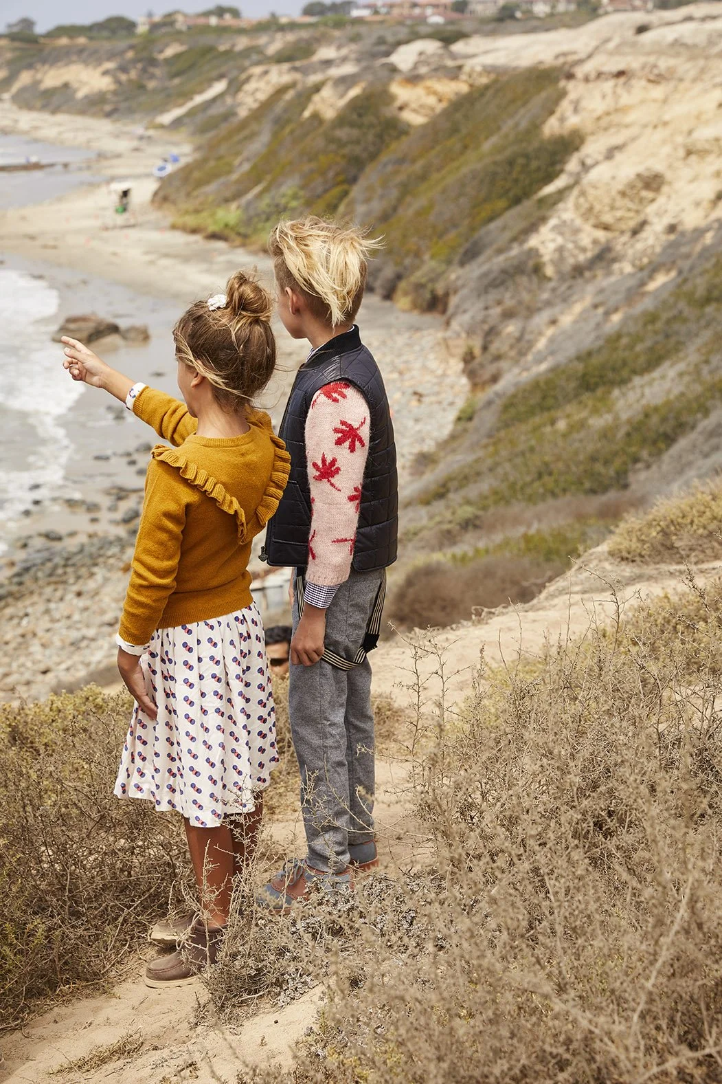 A girl and boy stand on a sandy beach cliff, looking out at the ocean. The girl points towards the water, wearing a yellow sweater and a white skirt with colorful dots. The boy, with tousled blonde hair, wears a black vest over a sweater with red flo