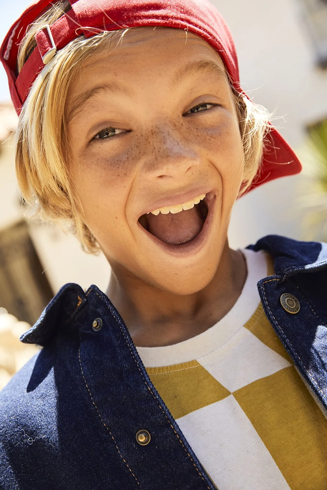 A young boy with blonde hair, freckles, and a red baseball cap, wearing a denim jacket over a yellow and white checkered shirt, laughing outdoors.