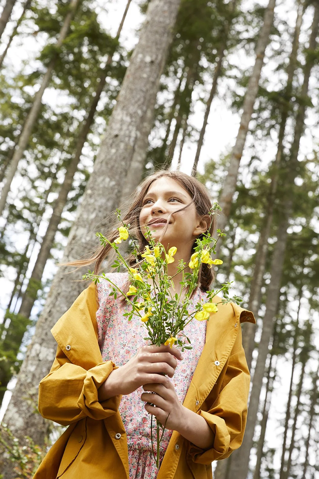 A woman in a yellow raincoat holding yellow flowers in a forest with tall trees and green foliage.