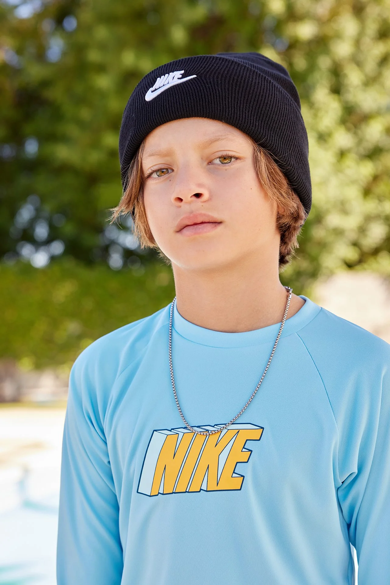 A teenage boy wearing a black Nike beanie, a sky blue Nike shirt, and a silver chain necklace, standing outdoors with blurred green trees in the background.