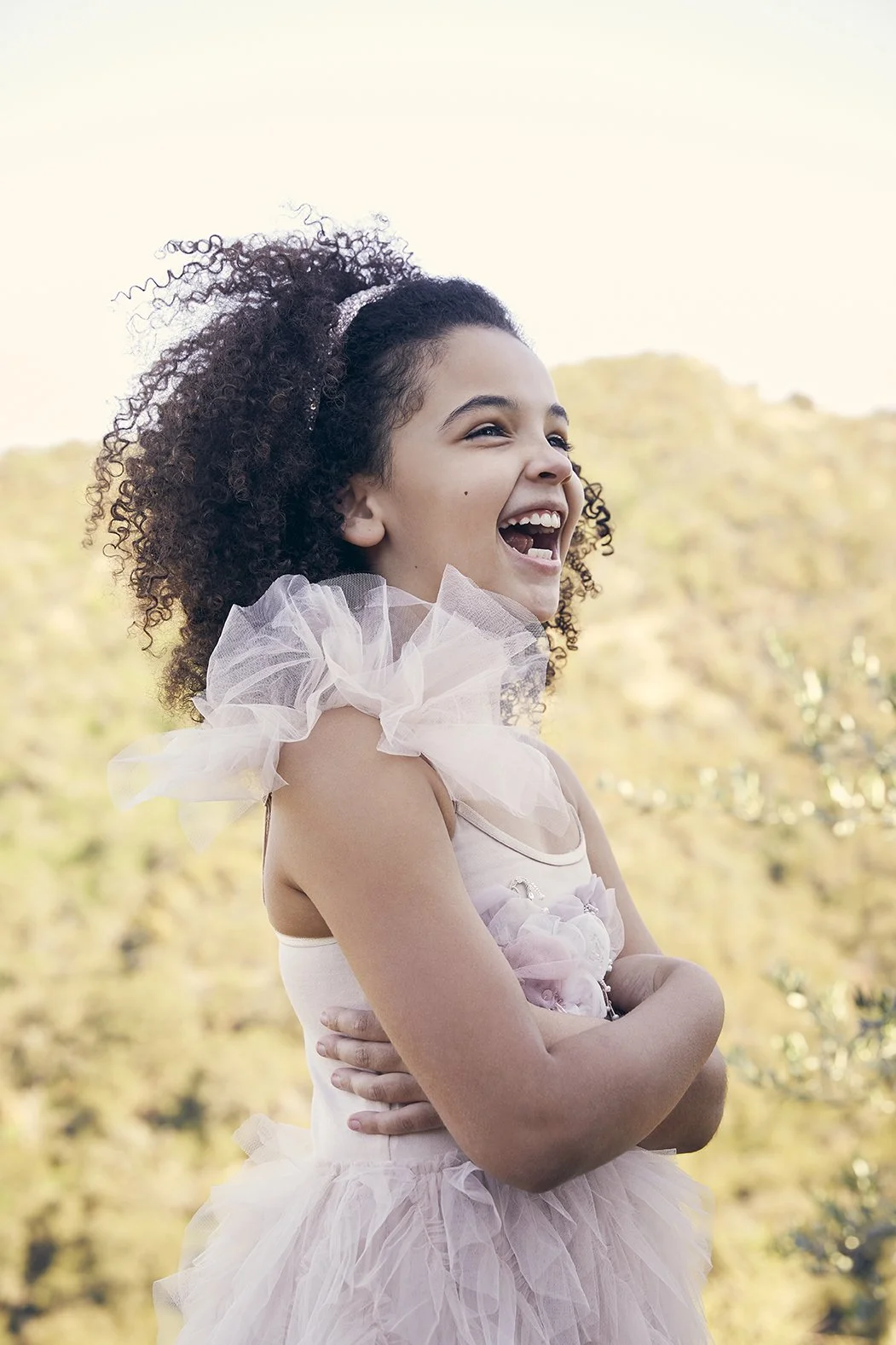 A young girl with curly hair, smiling and laughing outdoors, wearing a light pink dress with ruffled tulle sleeves.