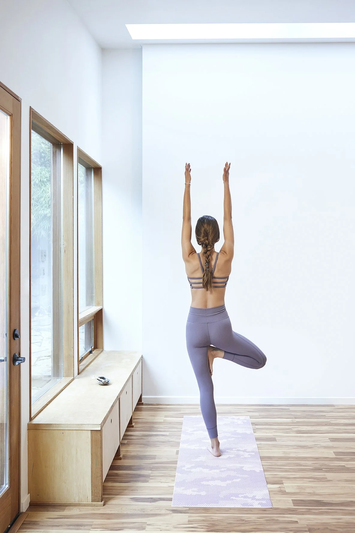 A woman practicing yoga indoors, performing the tree pose with arms extended upward and one foot resting on her inner thigh, on a yoga mat near large windows with natural light.