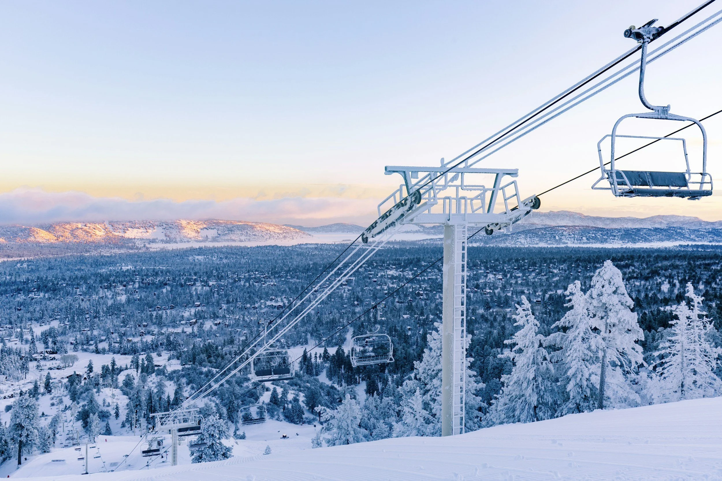 Empty ski lift chair with snow-covered landscape, trees, and distant mountains in the background during winter, with a clear sky.