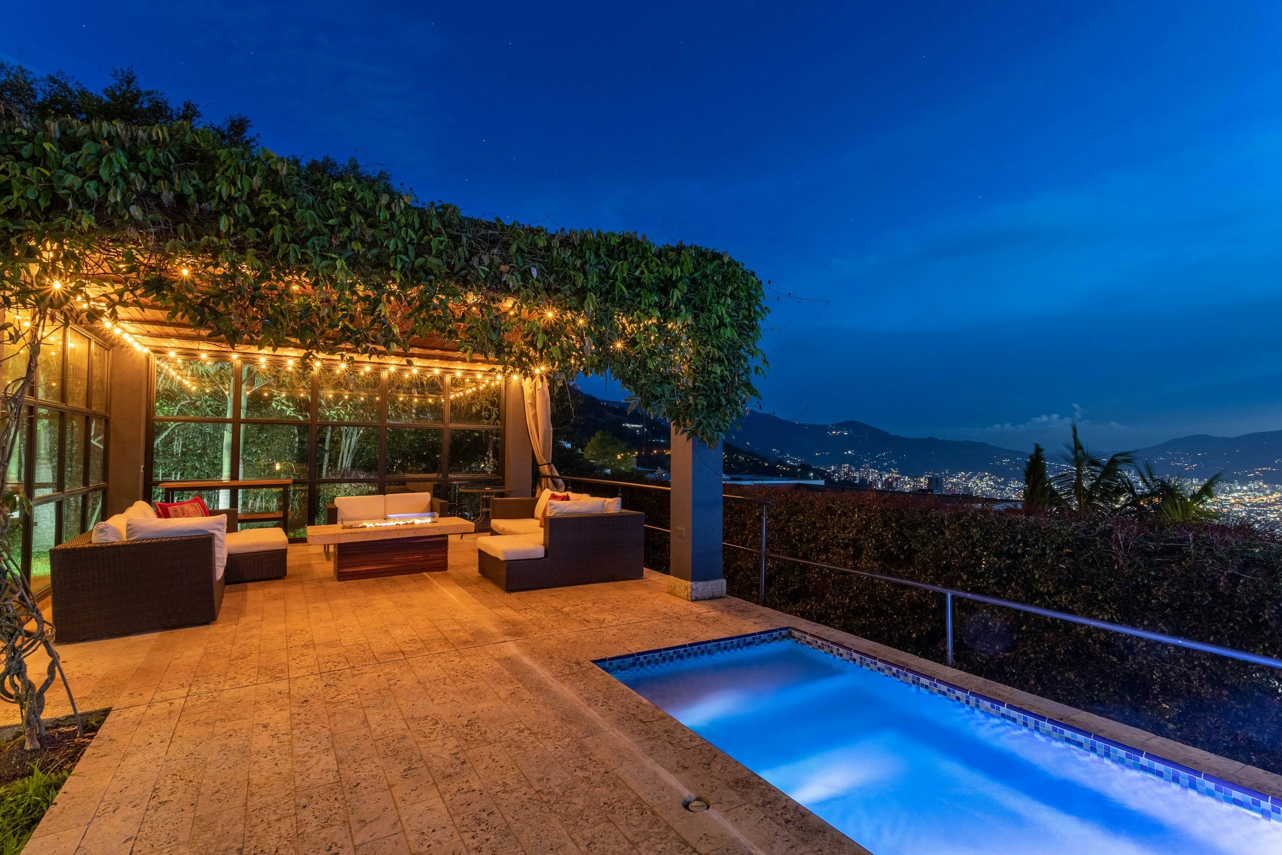 A nighttime view of a villa patio with outdoor seating, string lights, a small pool, overlooking a cityscape and mountains.