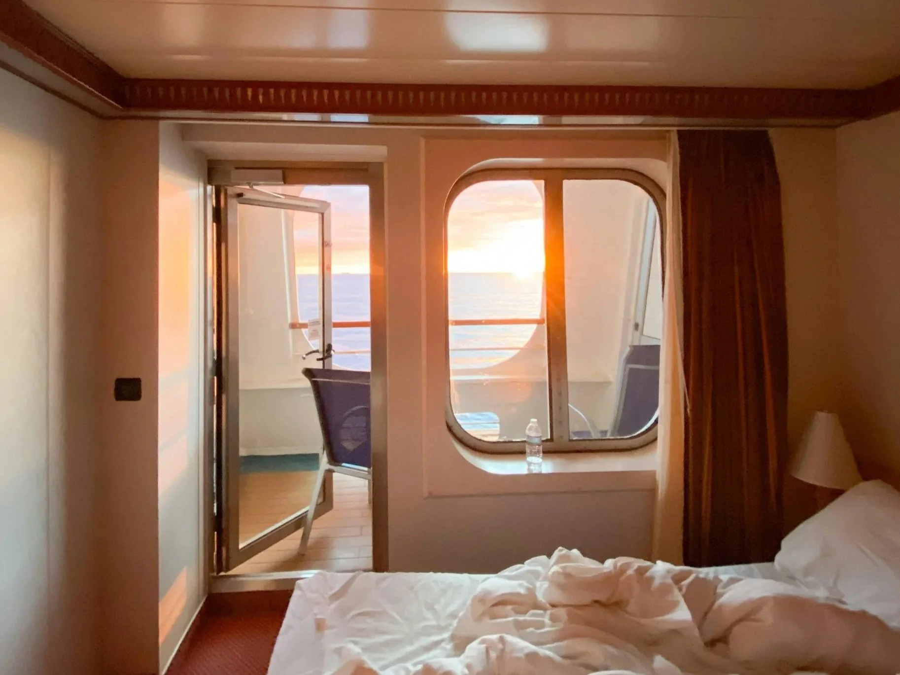 Inside a cruise ship cabin at sunset, showing a bed with rumpled white sheets, a chair, an open door leading to a balcony with a view of the ocean, and warm, ambient lighting.