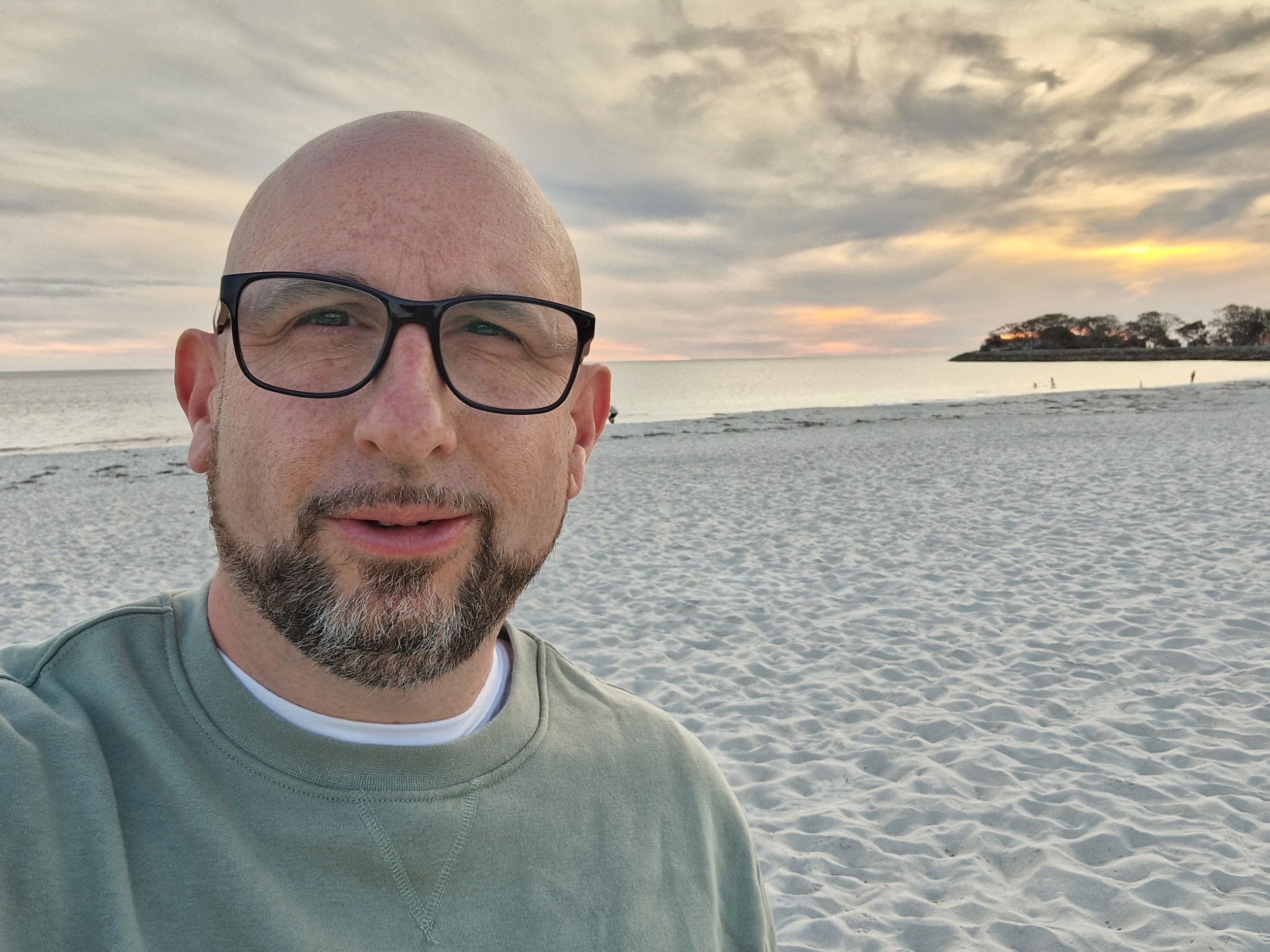 A man with glasses and a beard taking a selfie on a beach at sunset.