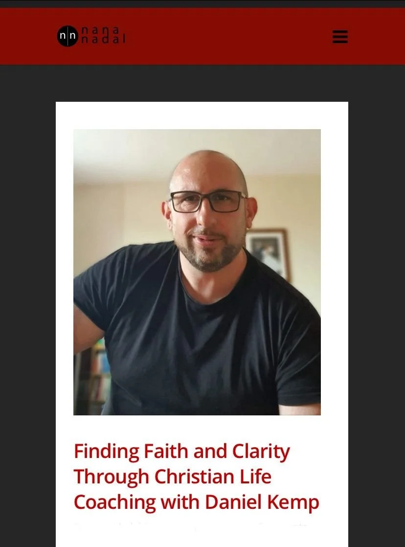 A man with glasses and a beard wearing a black shirt, smiling and sitting indoors with a framed picture on the wall in the background.