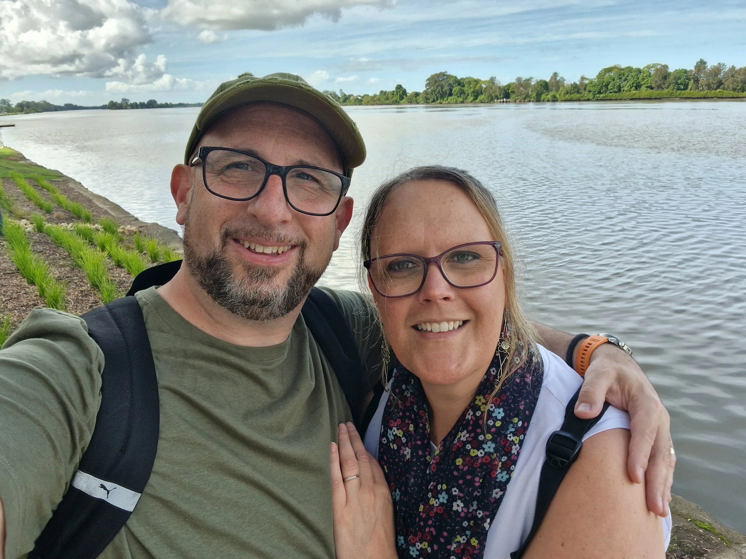 A smiling man and woman taking a close-up selfie by a river with green trees in the background. The man is wearing glasses, a hat, a green shirt, and a backpack. The woman has glasses, shoulder-length hair, and is wearing a white top with a floral scarf and a backpack.