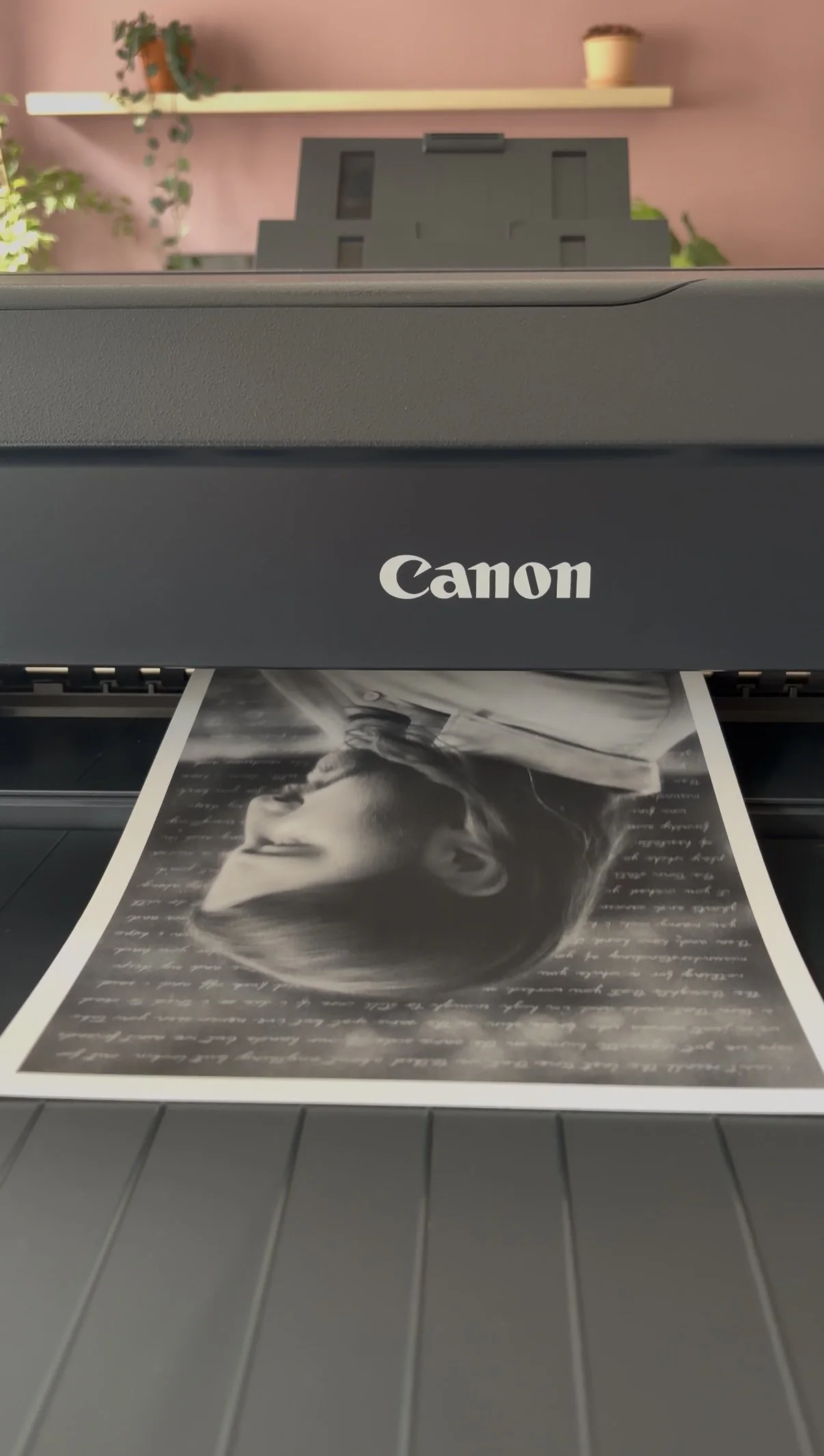 A Canon printer producing a black and white photo of a woman with glasses, with a pink wall, a wooden shelf with potted plants, and a beige pot in the background.