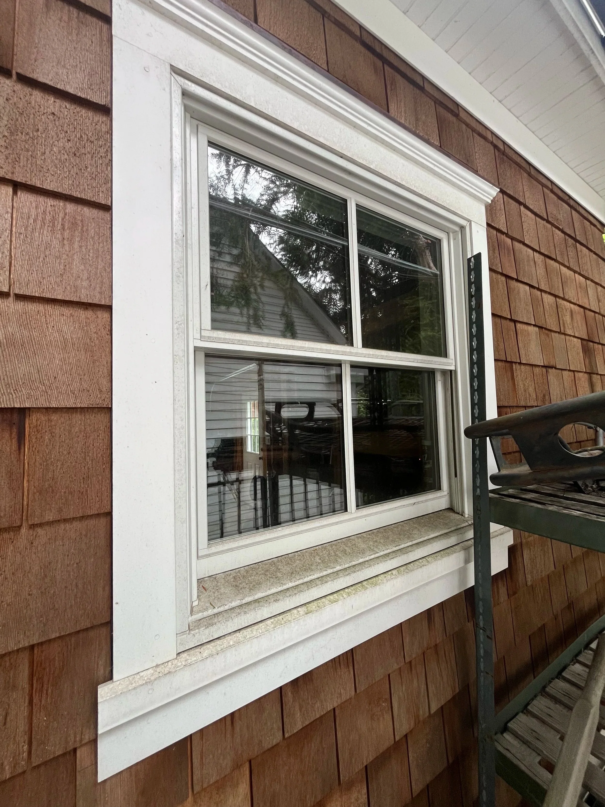Close-up of a window with a white frame on a house with brown shingle siding. The window has four panes with black screens. There is a metal support bracket on the right side of the window and a wooden bench or chair on the right side of the image.