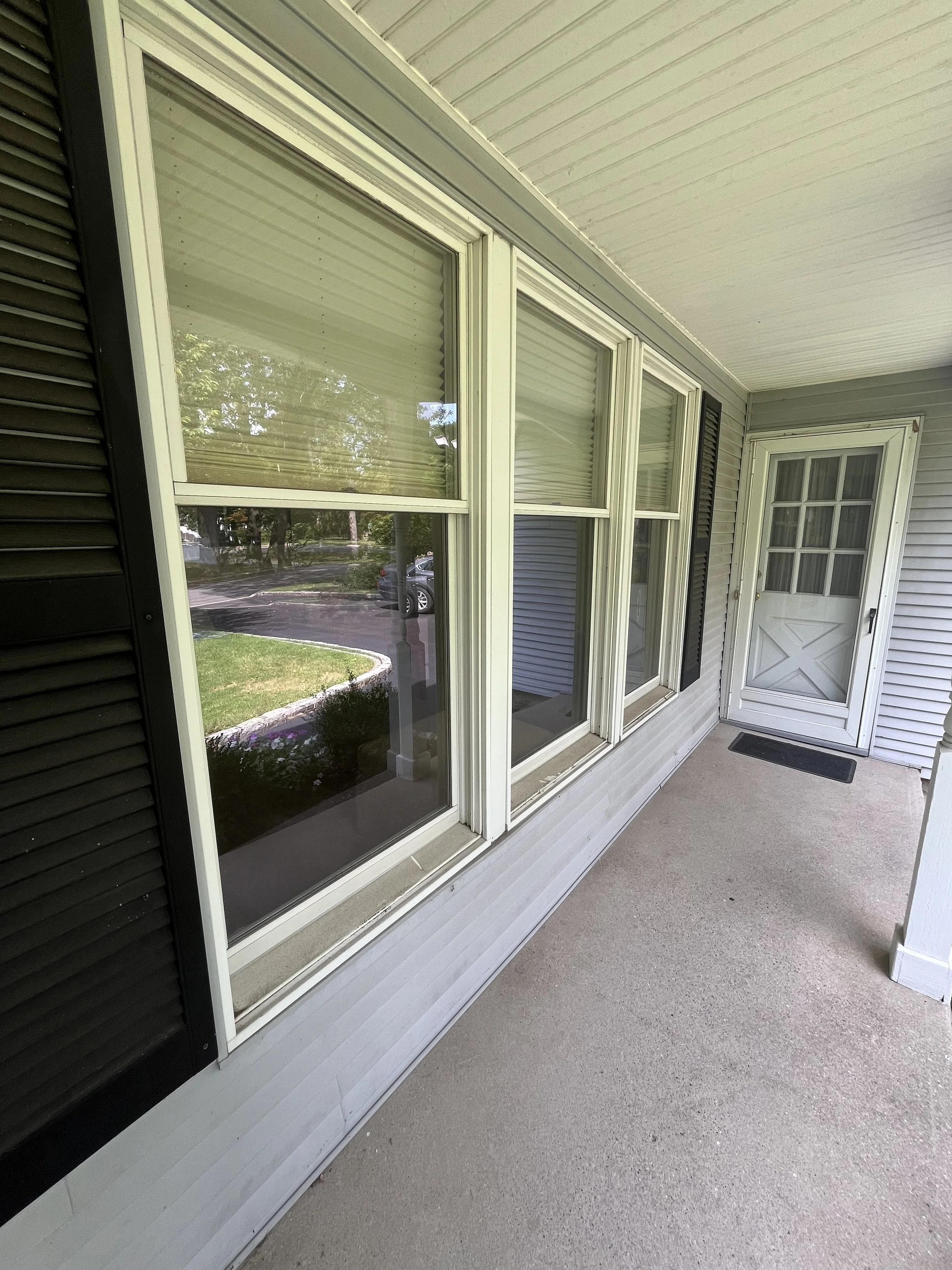 View of a front porch with four large windows with white frames, black shutters on the sides, and a door with a small window. The porch has a concrete floor and is part of a house with light gray siding. The reflection in the windows shows trees, a c