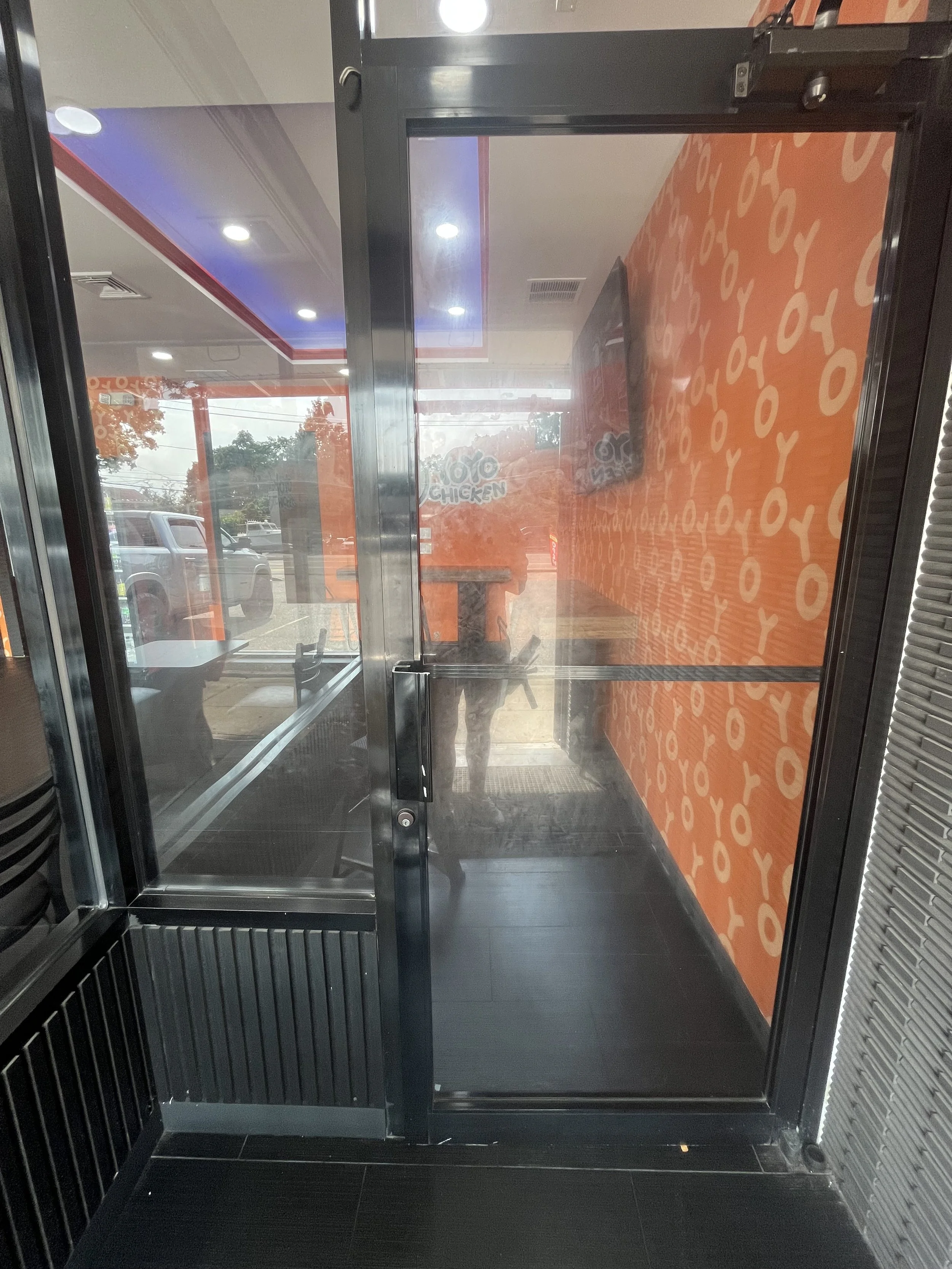 View through glass door into a fast food restaurant with black flooring, orange wall with patterned design, and a TV mounted on the wall. Outside the restaurant, there are parked cars and trees visible through the glass.