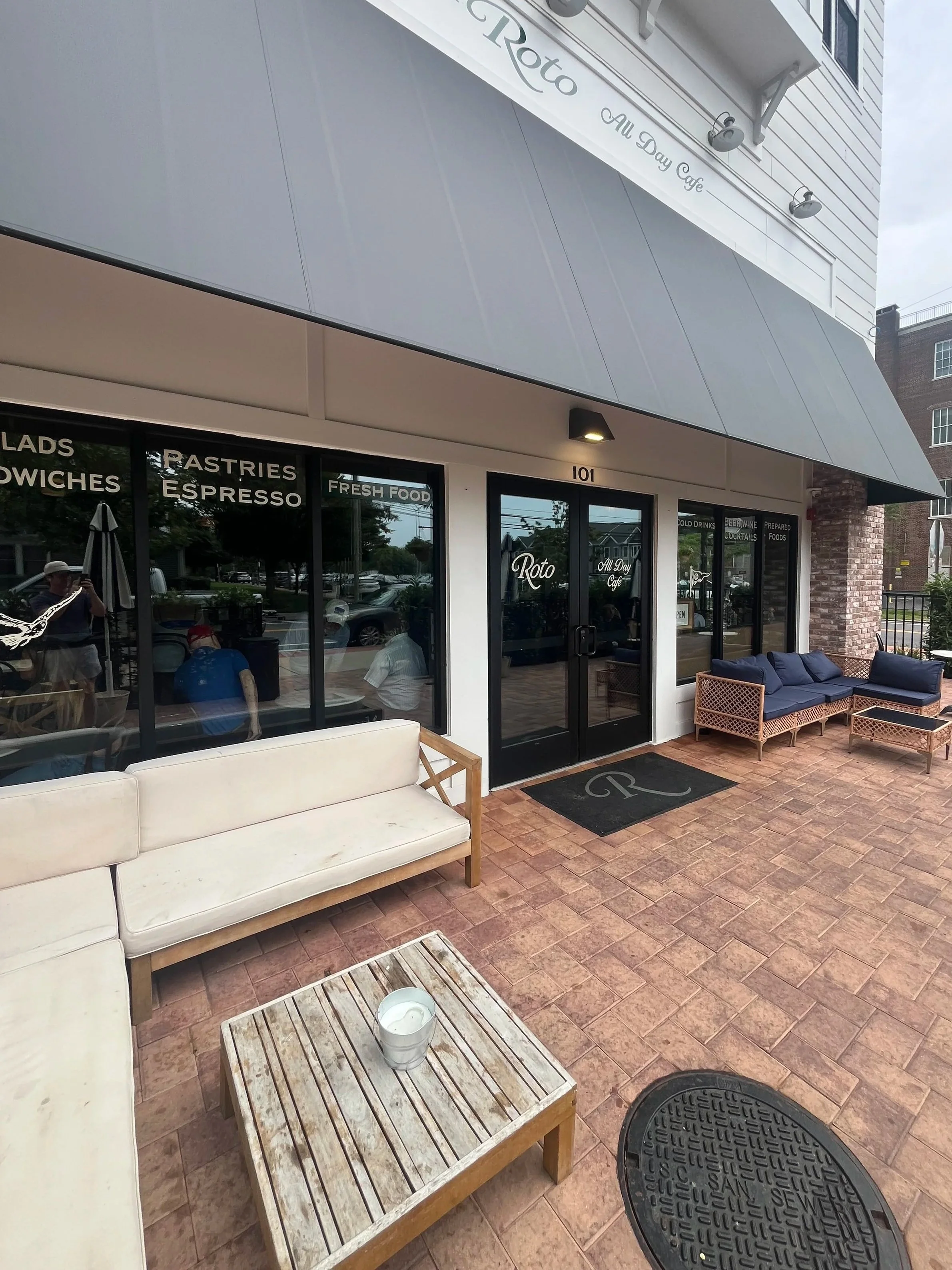 Exterior view of a cafe with a sliding glass door entrance, outdoor seating with a white couch, a wooden coffee table with a white candle, blue outdoor sofa, and brick patio. The cafe is named 'Roto All Day Cafe', located at 101, with signage on the door and window indicating food and drink offerings.