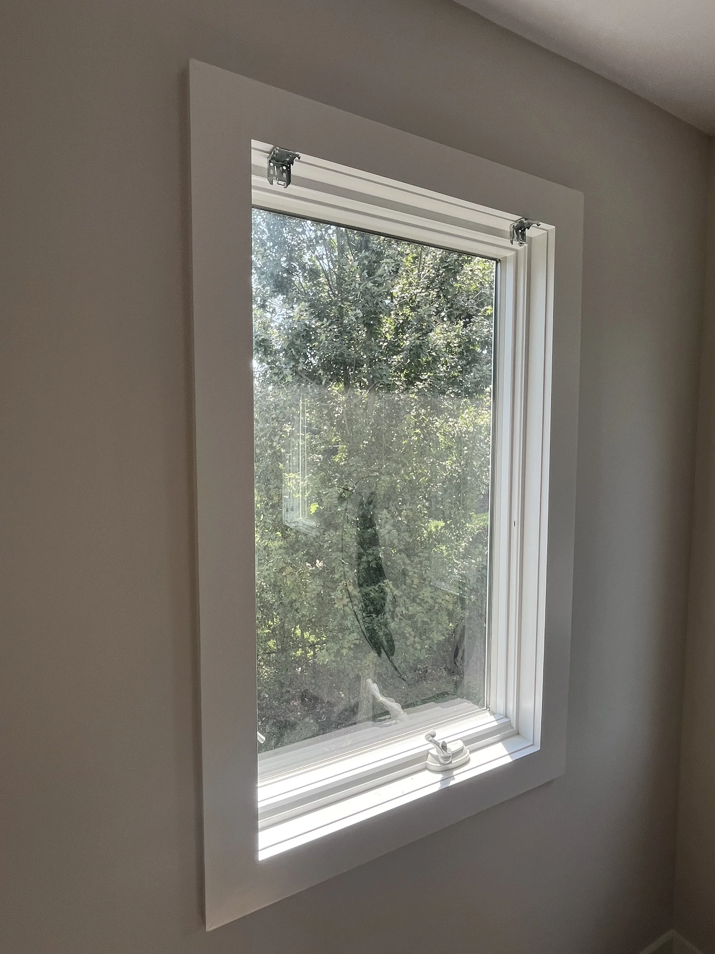 A white-framed window partially open with trees visible outside, situated in a room with beige walls.