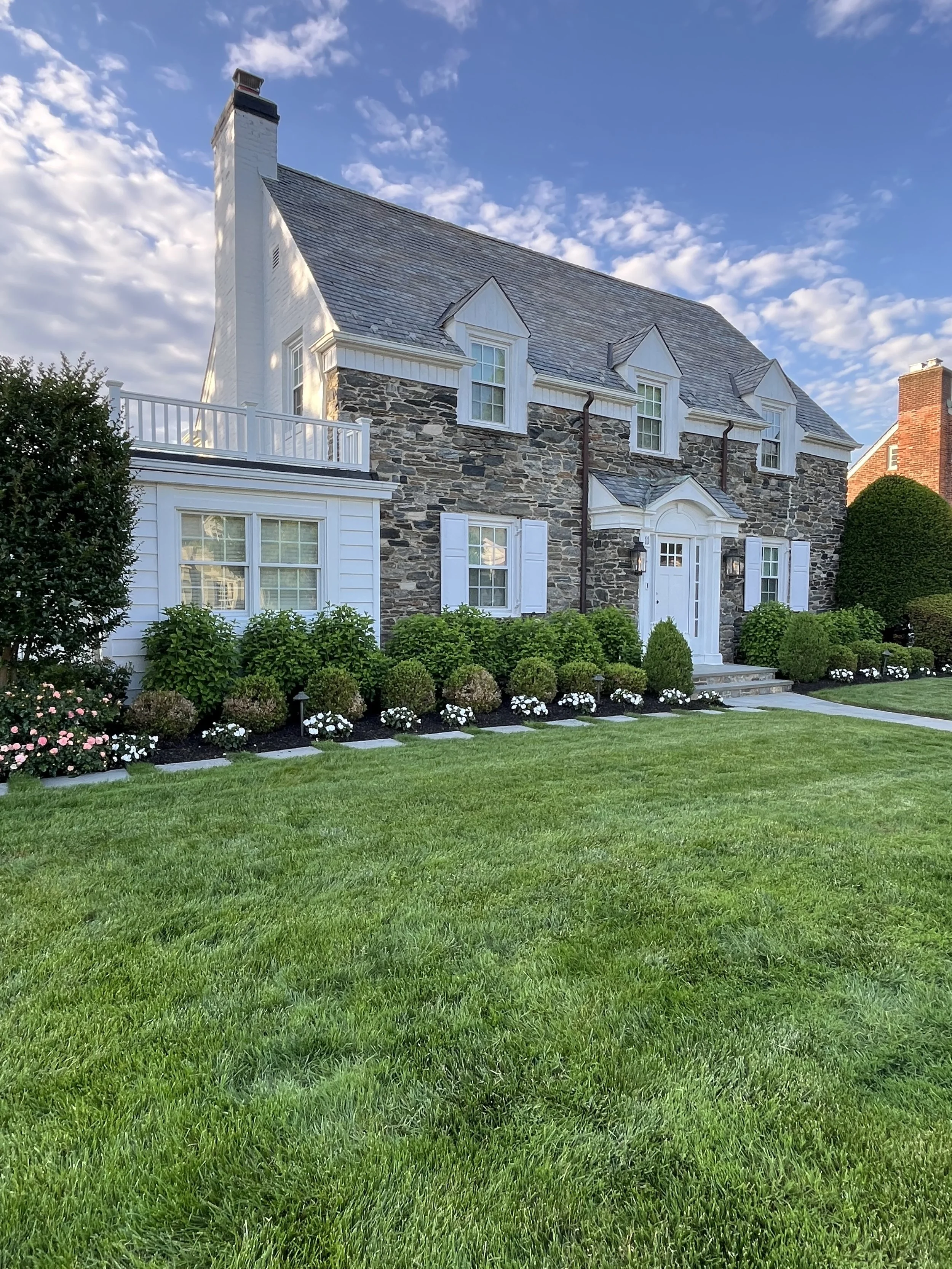 A large house with stone and white siding exterior, multiple dormer windows, a front staircase, surrounded by well-manicured green lawn and shrubbery, under a bright blue sky with scattered clouds.