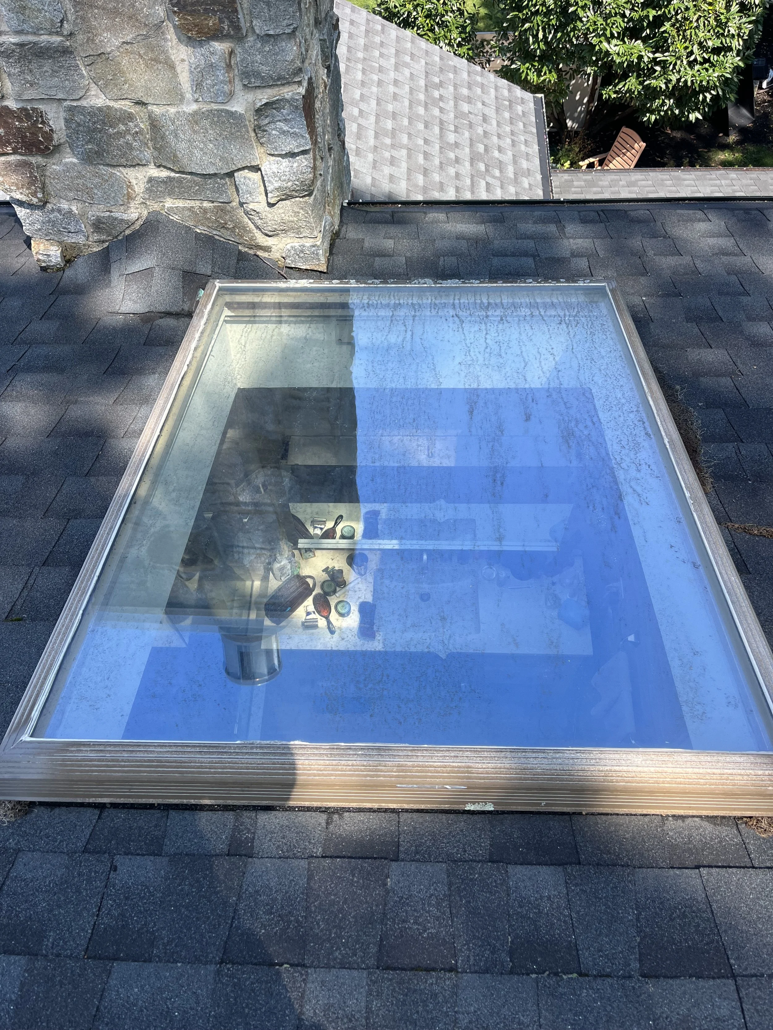 View of a rooftop with a large skylight window, black asphalt shingles, a stone chimney, and some trees in the background.