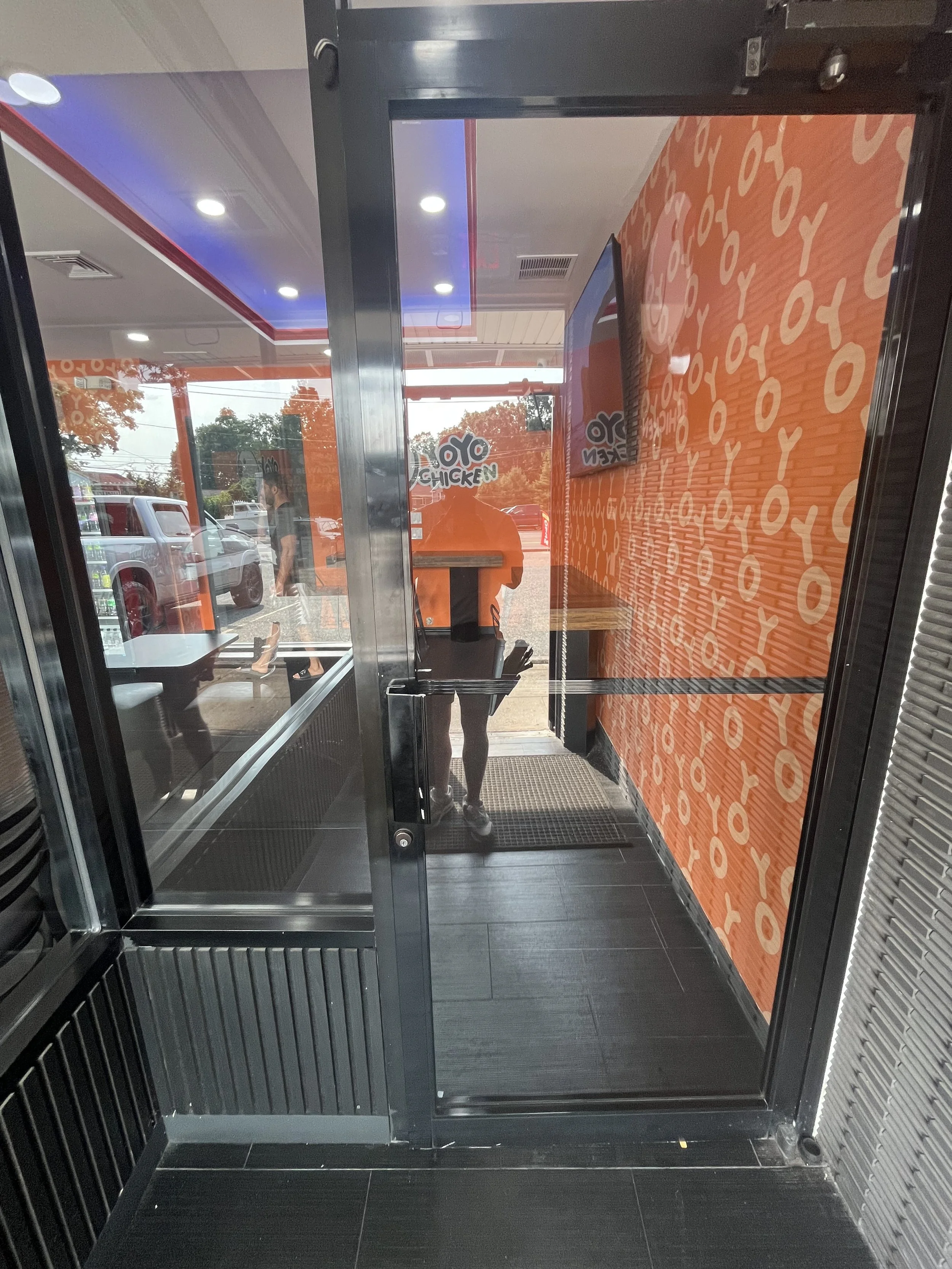 Inside a fast-food restaurant with orange and black decor, a person is walking out through a glass door, with several vehicles and trees visible outside.