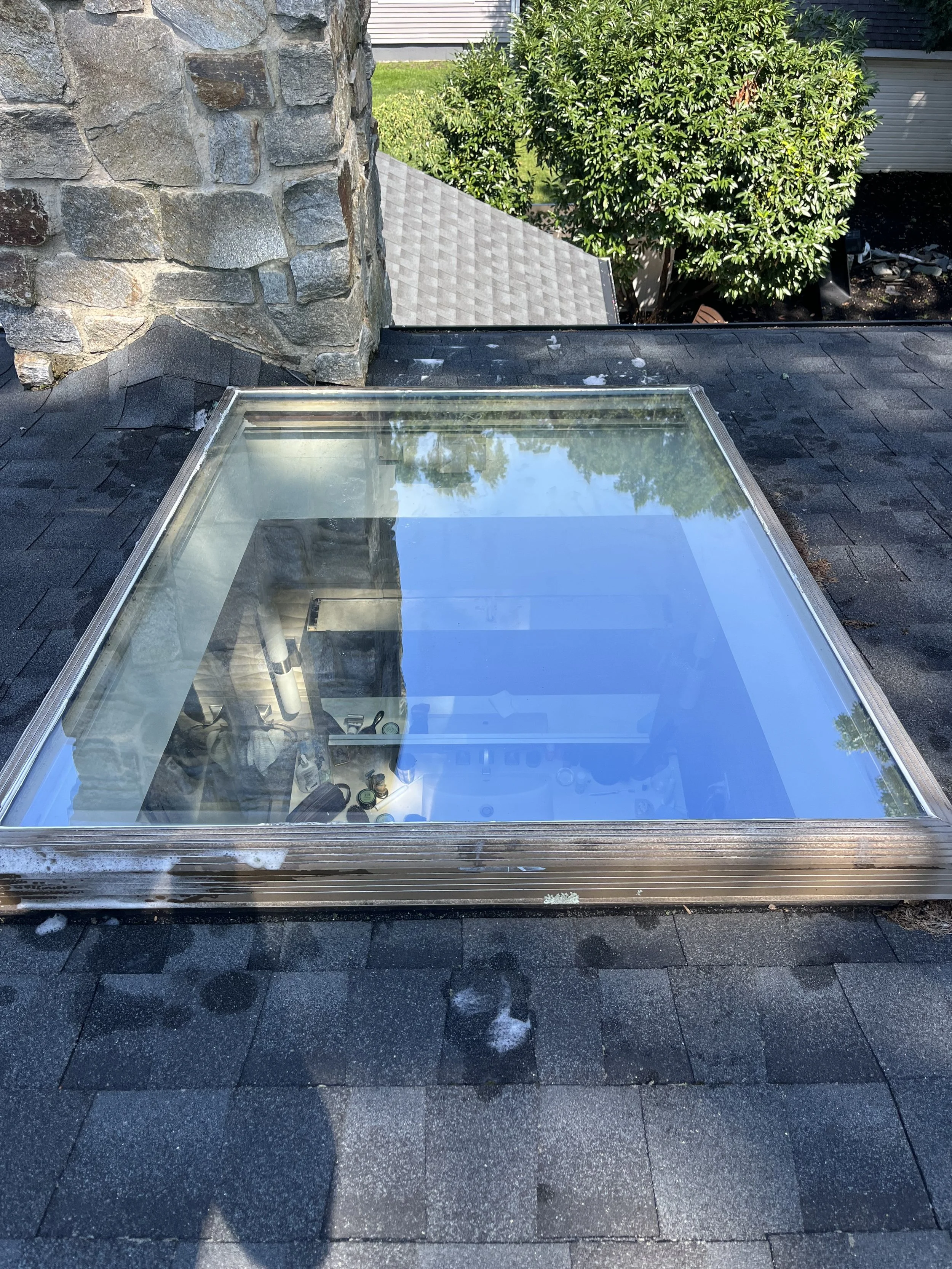 Top-down view of a glass skylight on a shingled roof, with a stone chimney on the left and green trees in the background.