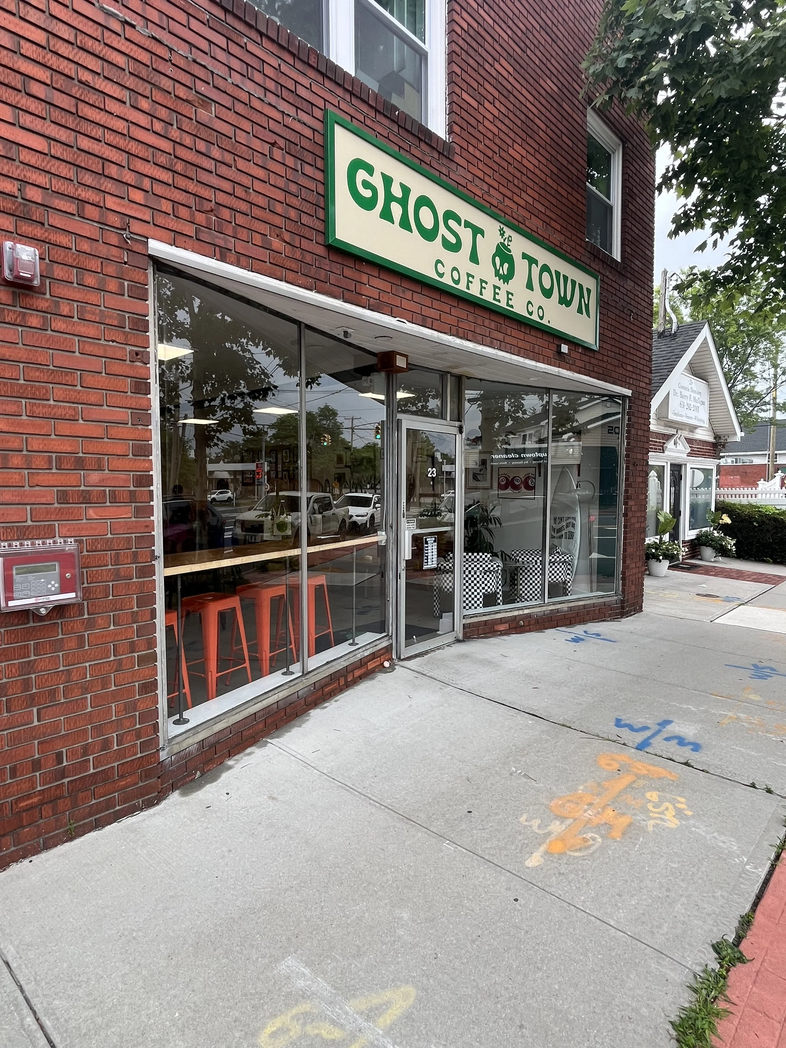 The exterior of a brick coffee shop named Ghost Town Coffee Co., with large glass windows, red barstools inside, and sidewalk parking lines. Ongoing sidewalk chalk art is visible on the pavement outside.