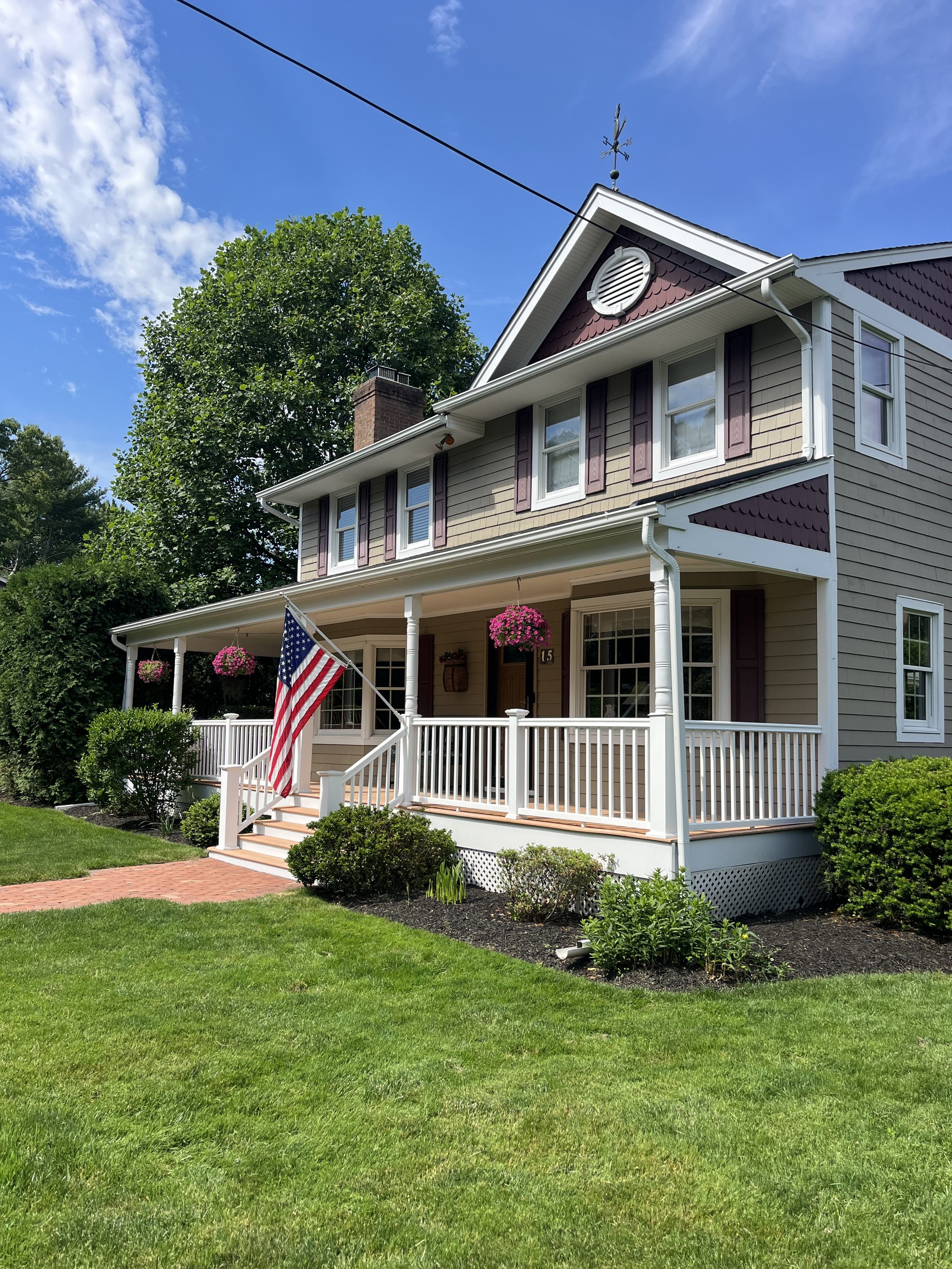A beige and purple house with a porch, American flag, hanging flower baskets, surrounded by a green lawn and bushes, under a blue sky with some clouds.
