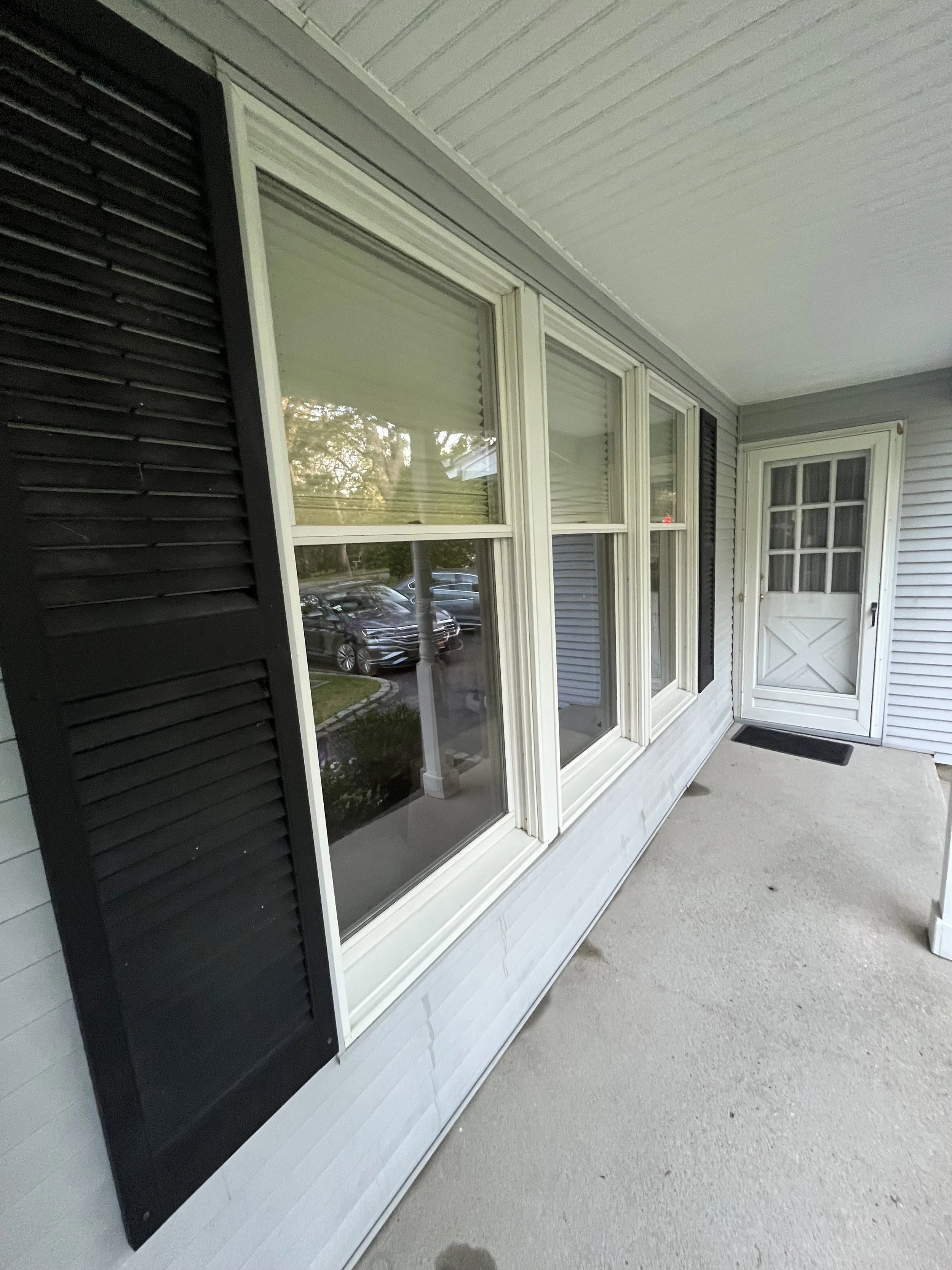 The image shows a window with white framing, black shutters on either side, and a white wooden door with a small window at the top, all on a porch with a concrete floor.