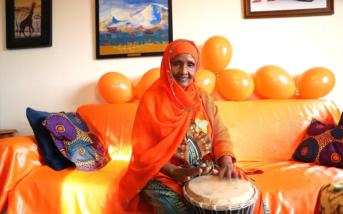 A woman dressed in orange with a headscarf sitting on an orange sofa, surrounded by orange balloons, playing a drum. The room has framed artwork on the wall behind her.