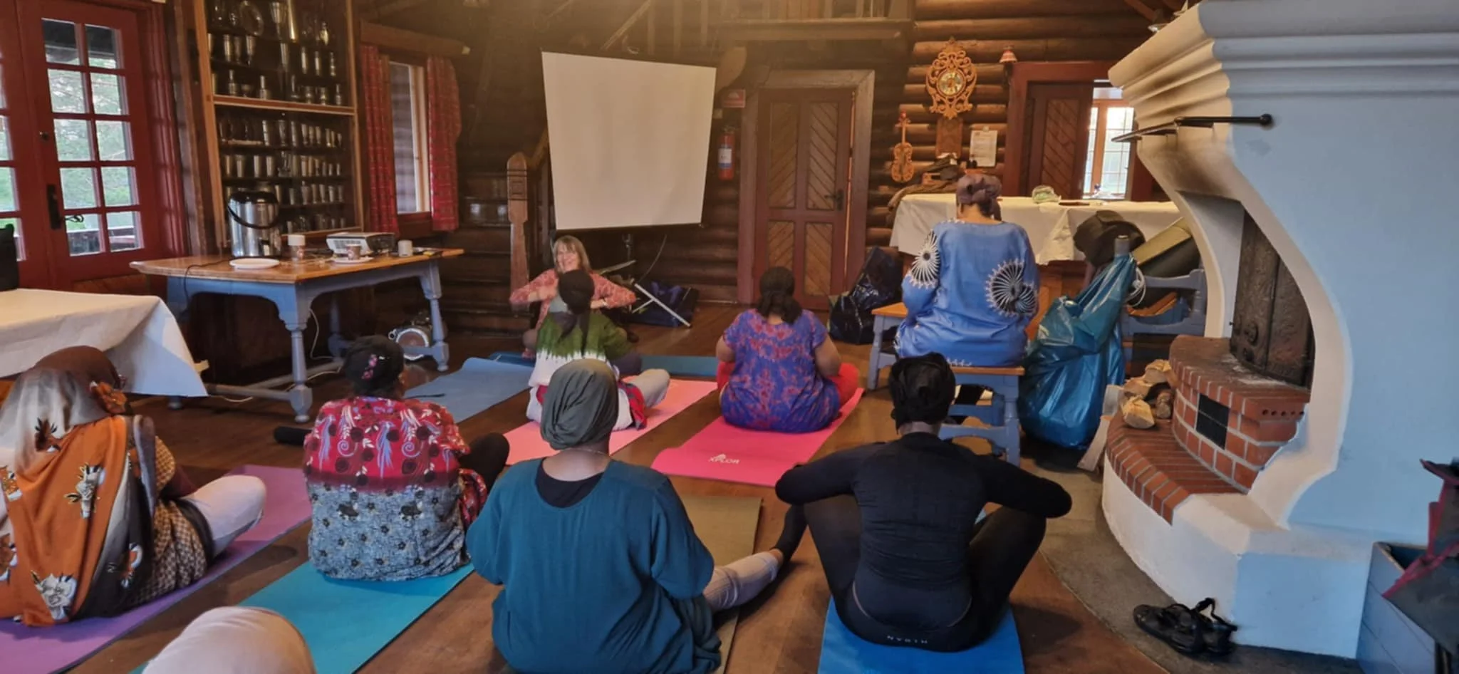 A group of people attending a yoga class inside a cozy wooden cabin, sitting on colorful yoga mats facing a instructor at the front near a white projection screen.