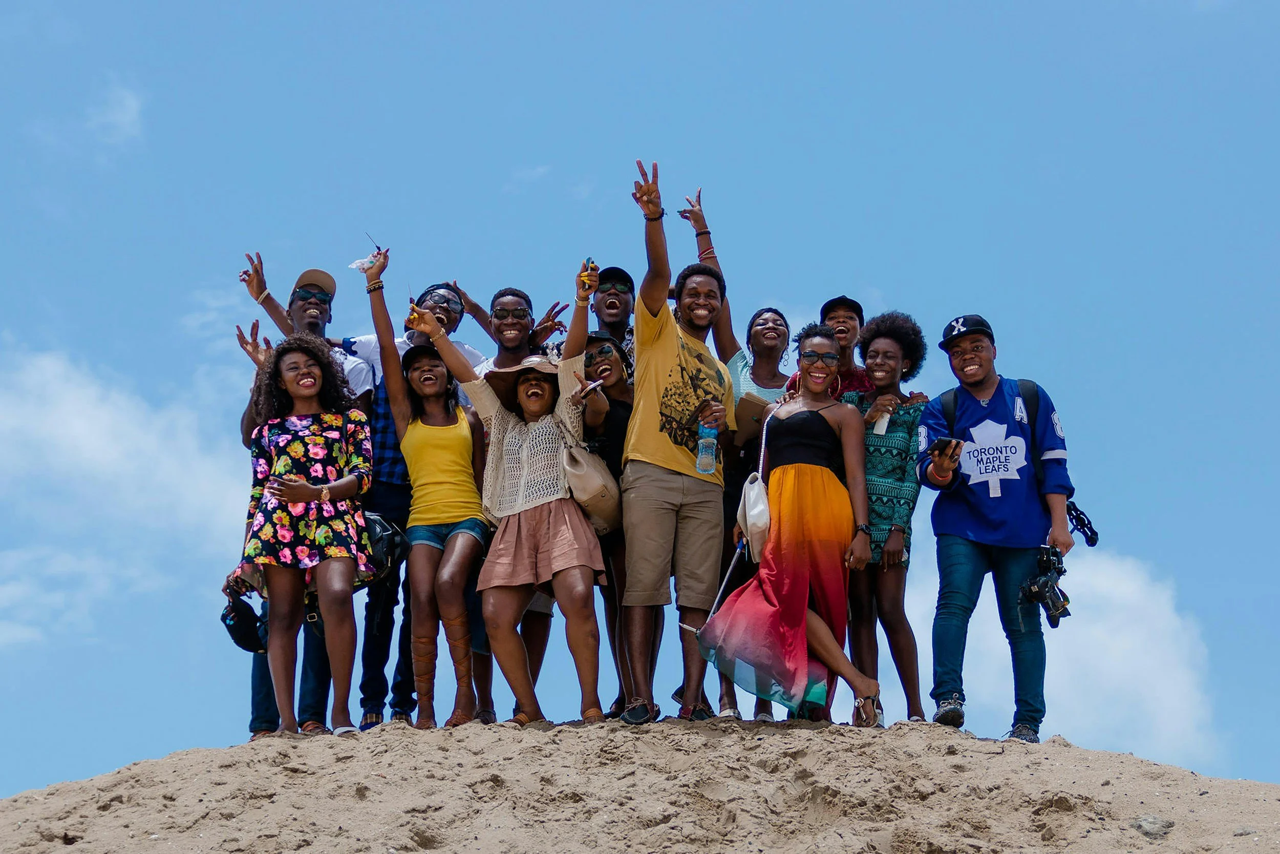 Group of people smiling and celebrating on a hilltop against a blue sky, some making peace signs and holding drinks.