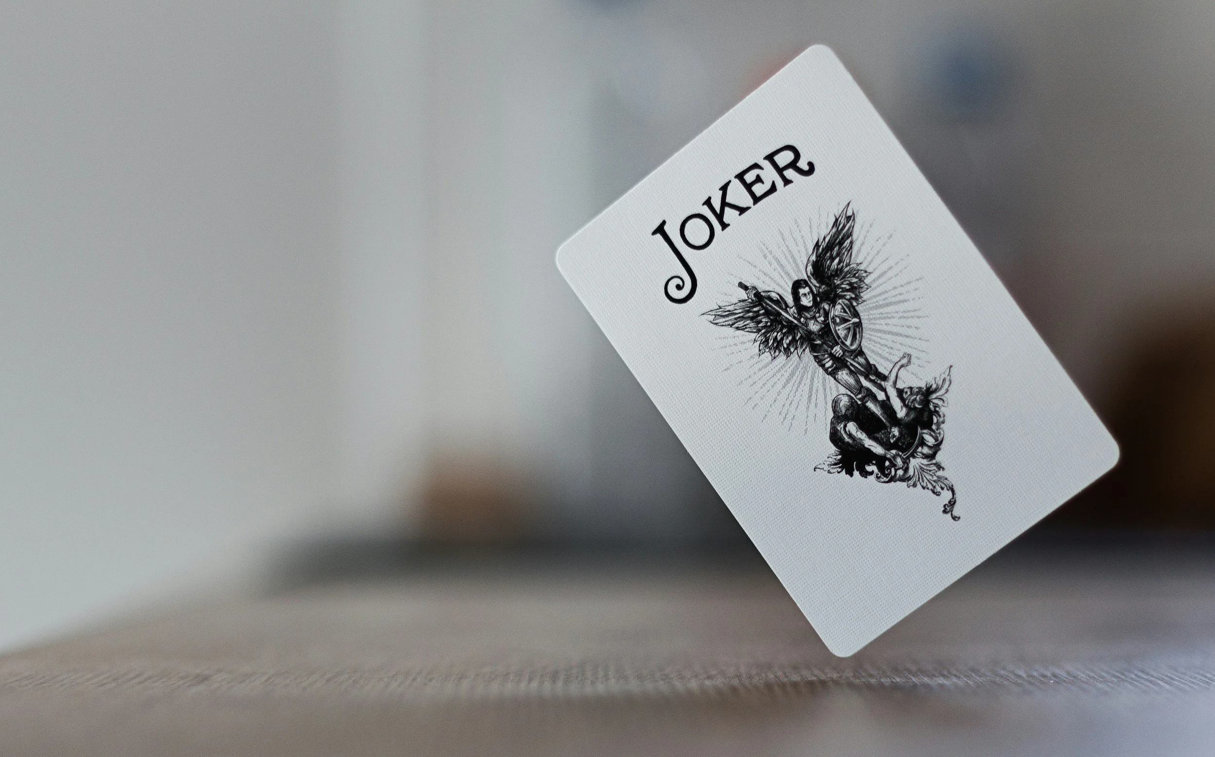 A Joker playing card hovering above a flat surface, showing an illustration of a winged woman with a shield and sword.