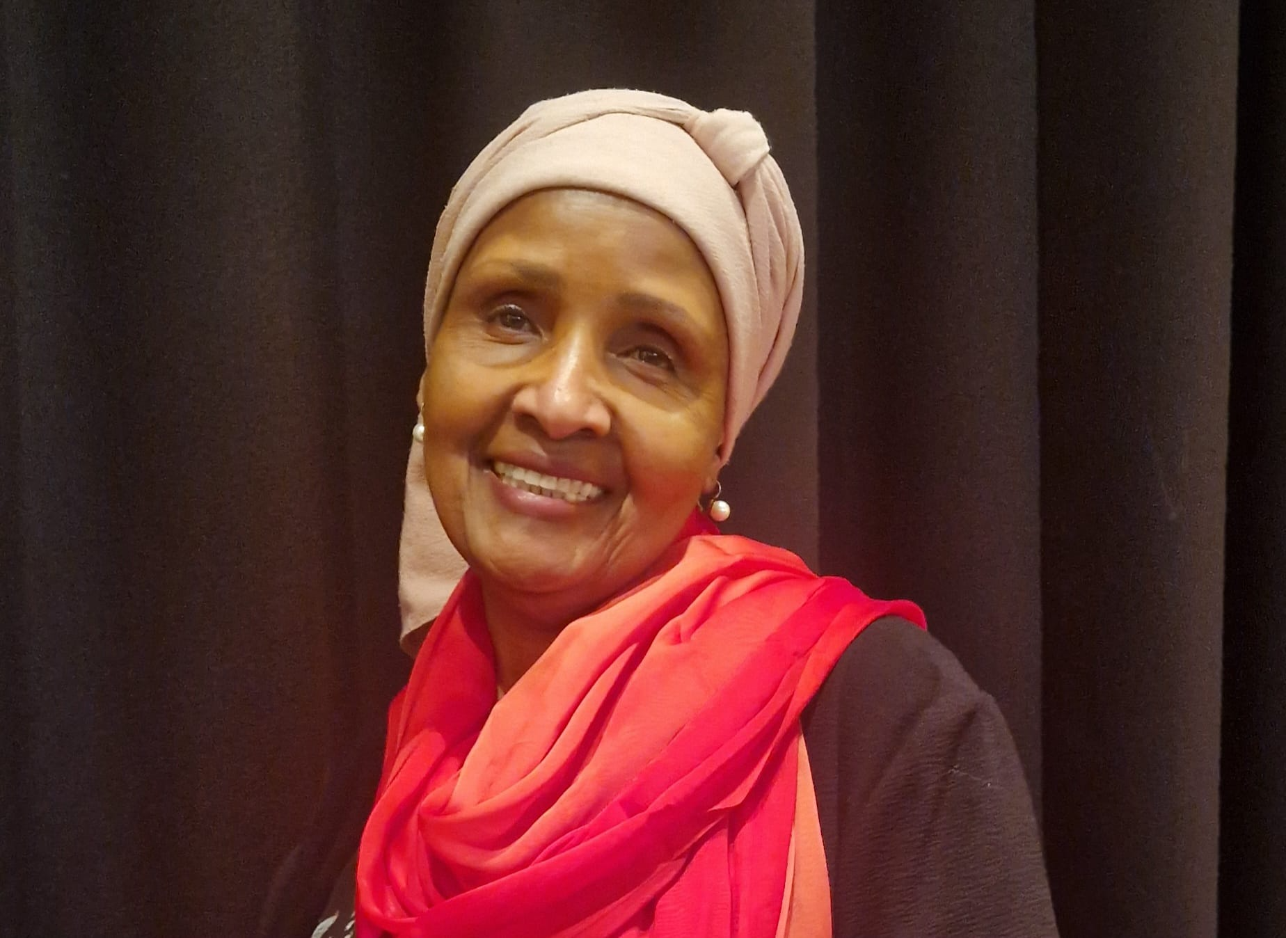 Smiling woman wearing a beige headwrap, black jacket, and red scarf, standing in front of black curtains.