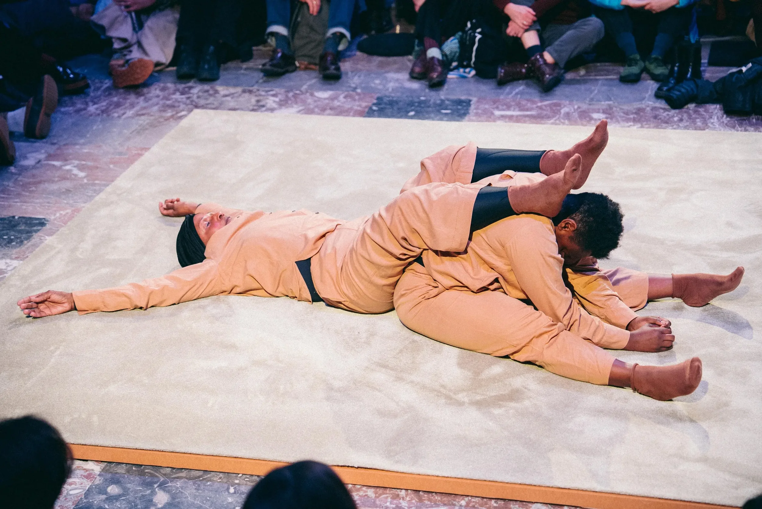 Two performers dressed in beige costumes are lying on a large beige mat, forming a human sculpture, with one performer on top of the other in a shared pose, while an audience watches in the background.