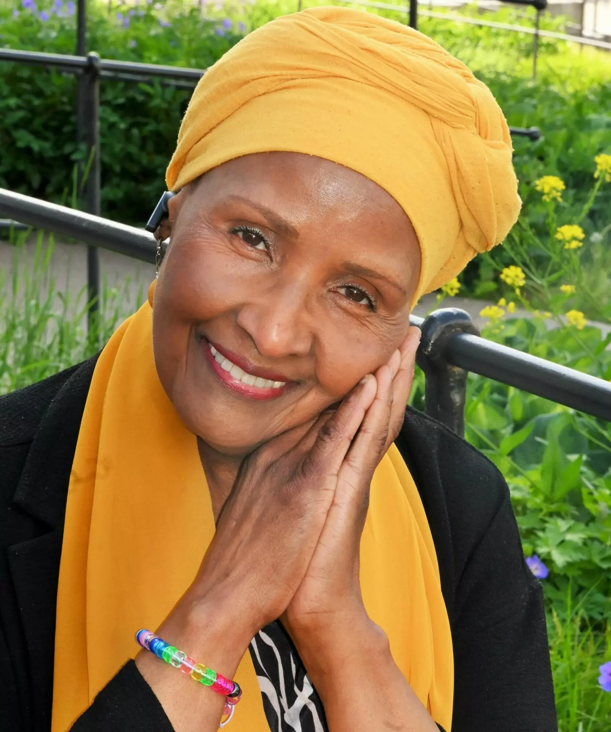 A woman smiling with her hand resting on her chin, wearing a yellow headwrap and scarf, outdoors with green foliage and purple flowers in the background.