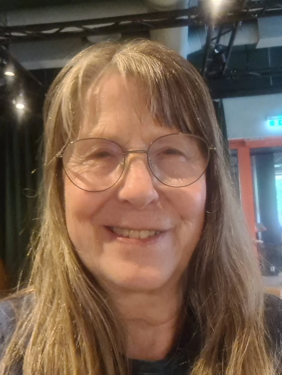 A smiling woman with long, light brown hair and glasses, indoors with a dark ceiling and a lit exit sign in the background.