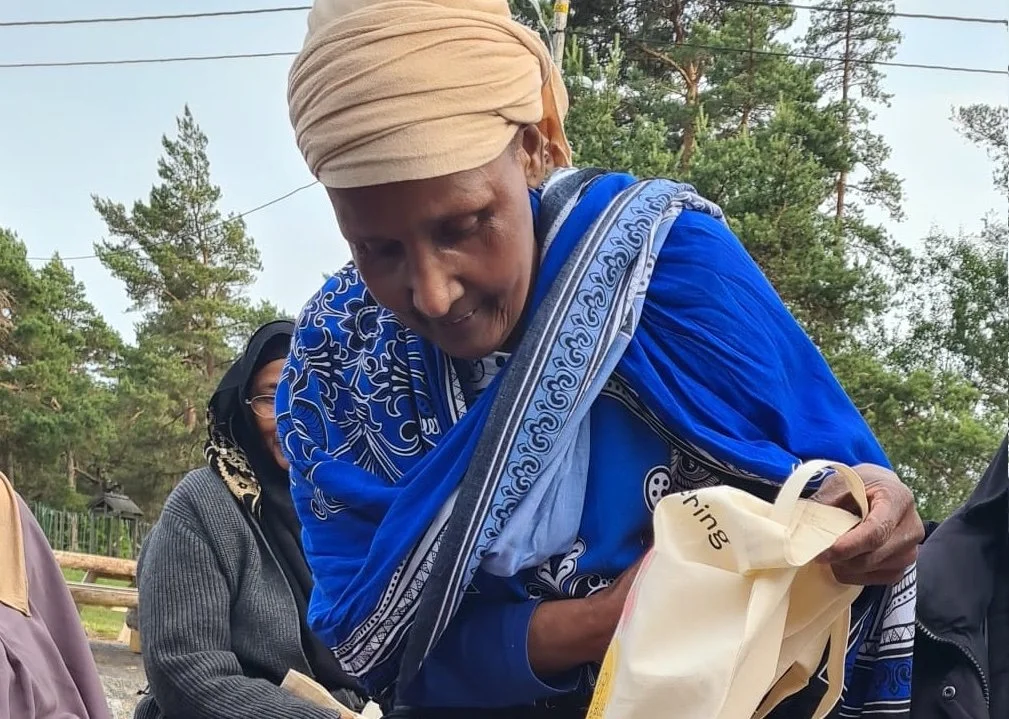 A woman in a beige headwrap and blue traditional attire looks into a bag, surrounded by other women outdoors in a wooded area.