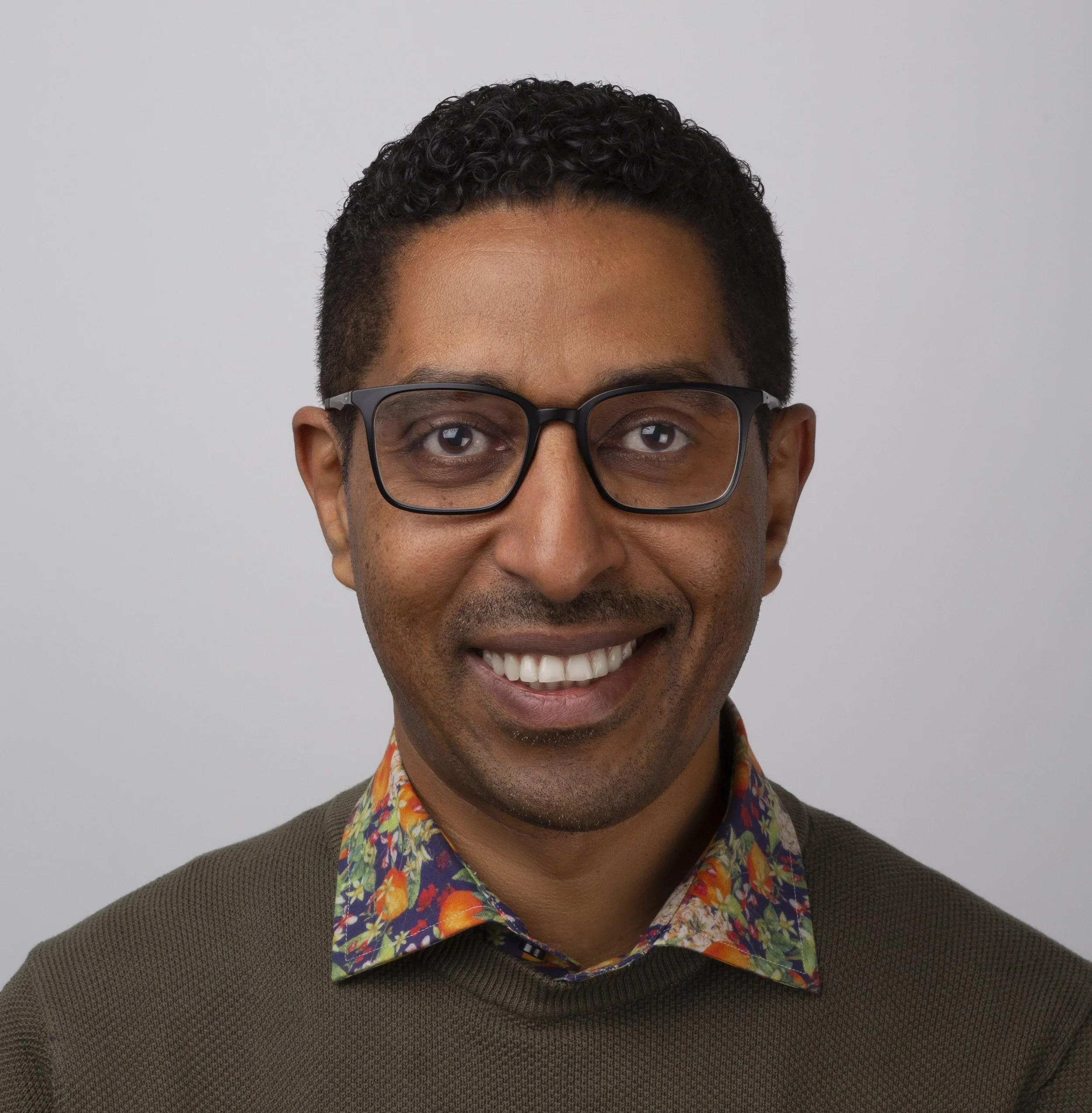 A man with curly dark hair, glasses, and wearing a dark shirt, smiling in front of a blue screen or background.