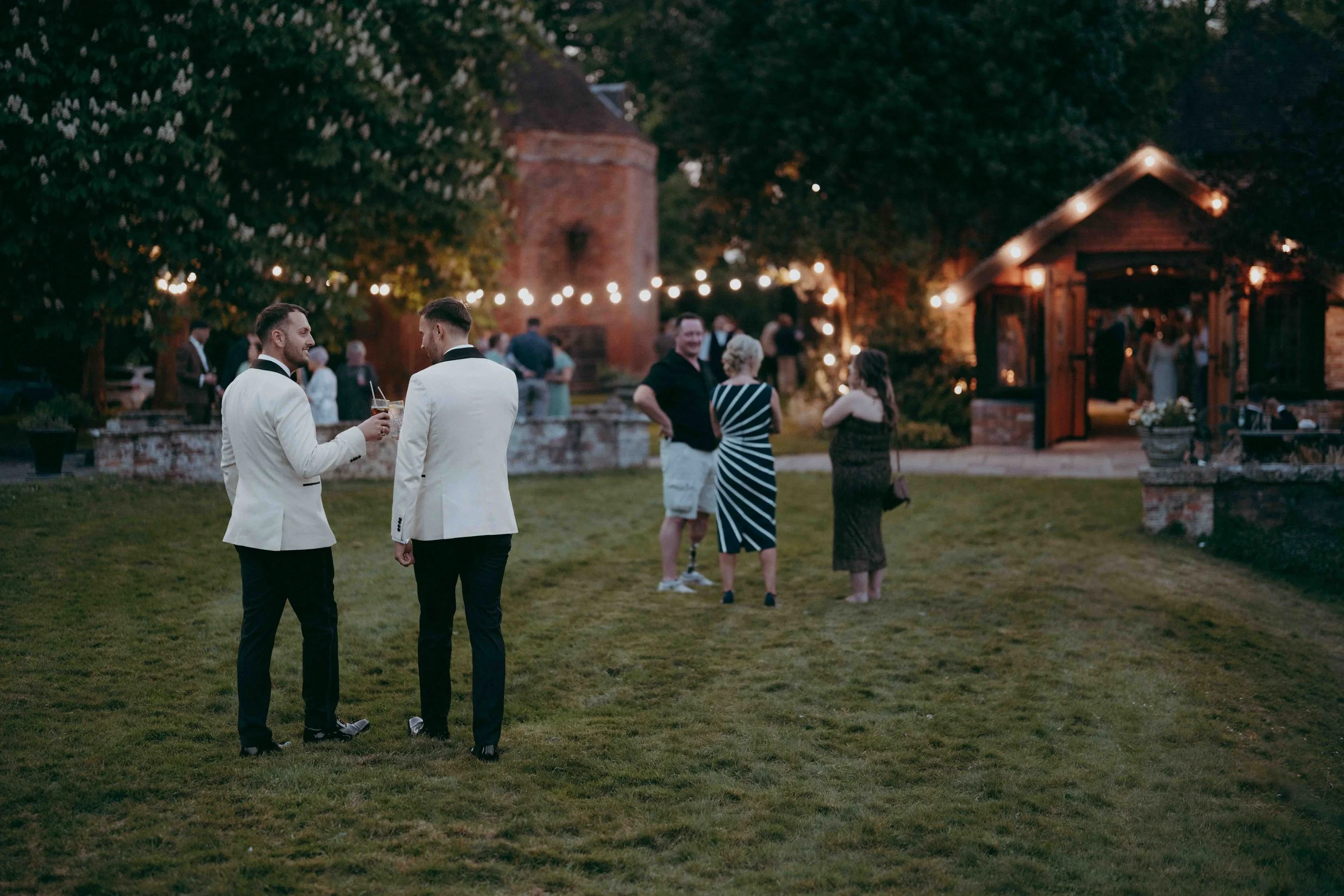 People enjoying an outdoor evening wedding reception with string lights, seated and standing in a garden area at Lainston House, near Winchester Hampshire, UK.