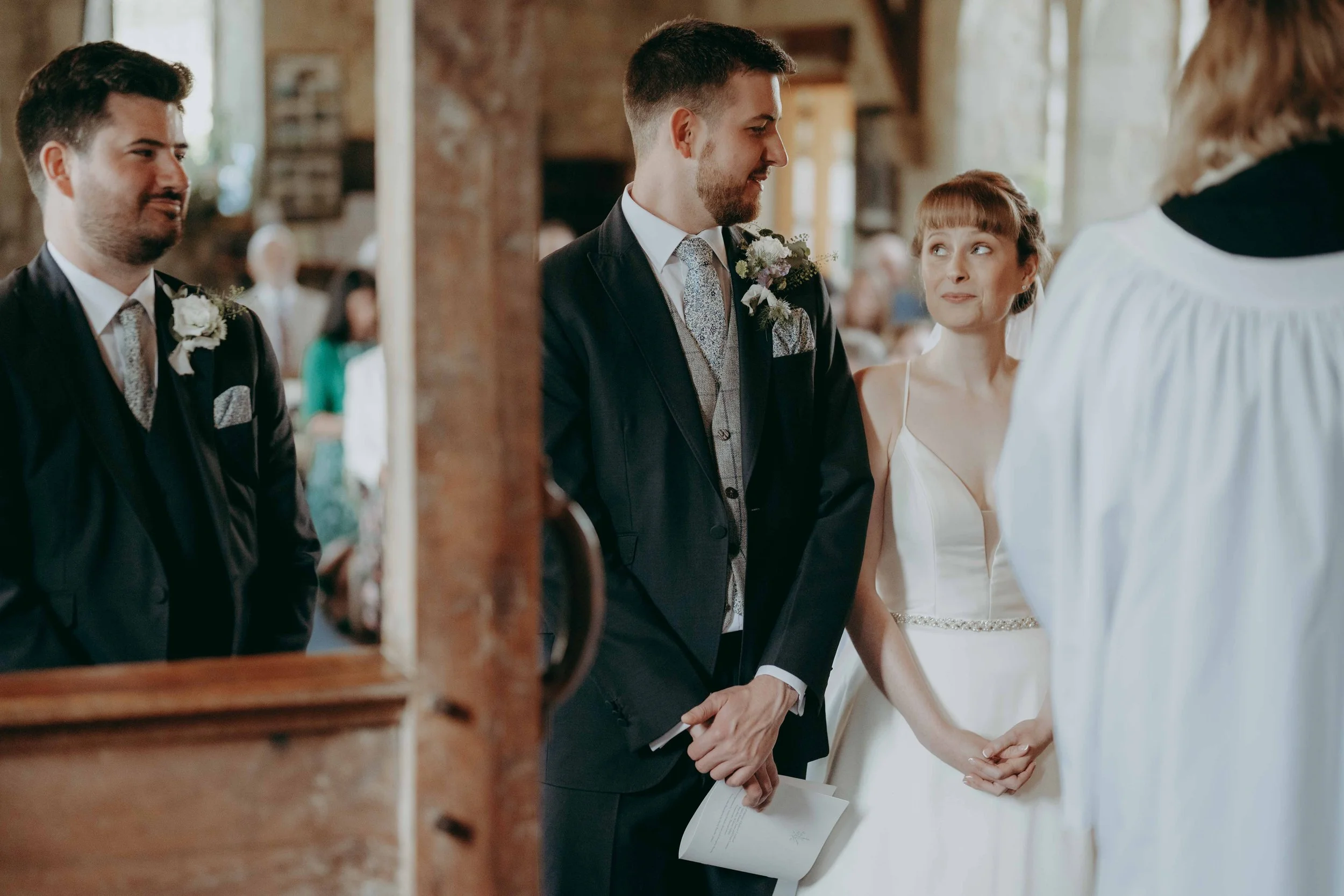 A couple getting married, holding hands, during a wedding ceremony inside a rustic venue. The bride is wearing a white gown, and the groom is in a dark suit. Taken at St Agnes Church, Freshwater, Isle of Wight, UK.