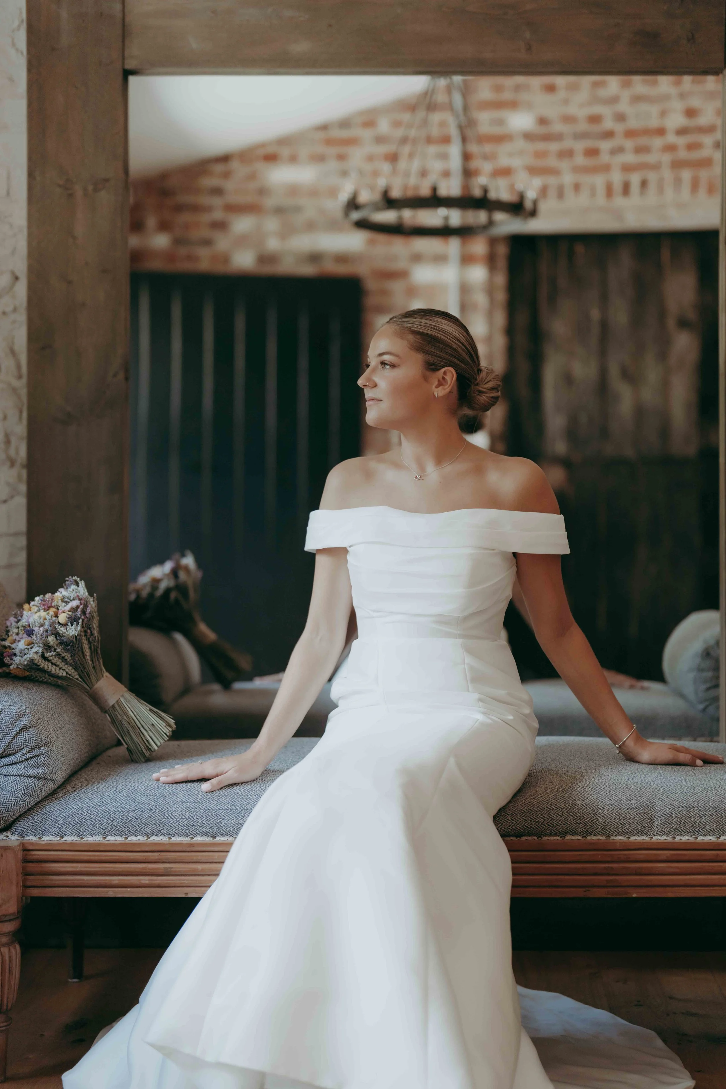 A bride at the Clock Barn wedding venue in Hampshire, UK, in a white wedding dress sitting on a cushioned window seat, looking to the left, with a bouquet of flowers beside her, in a rustic interior with brick walls and wooden beams.