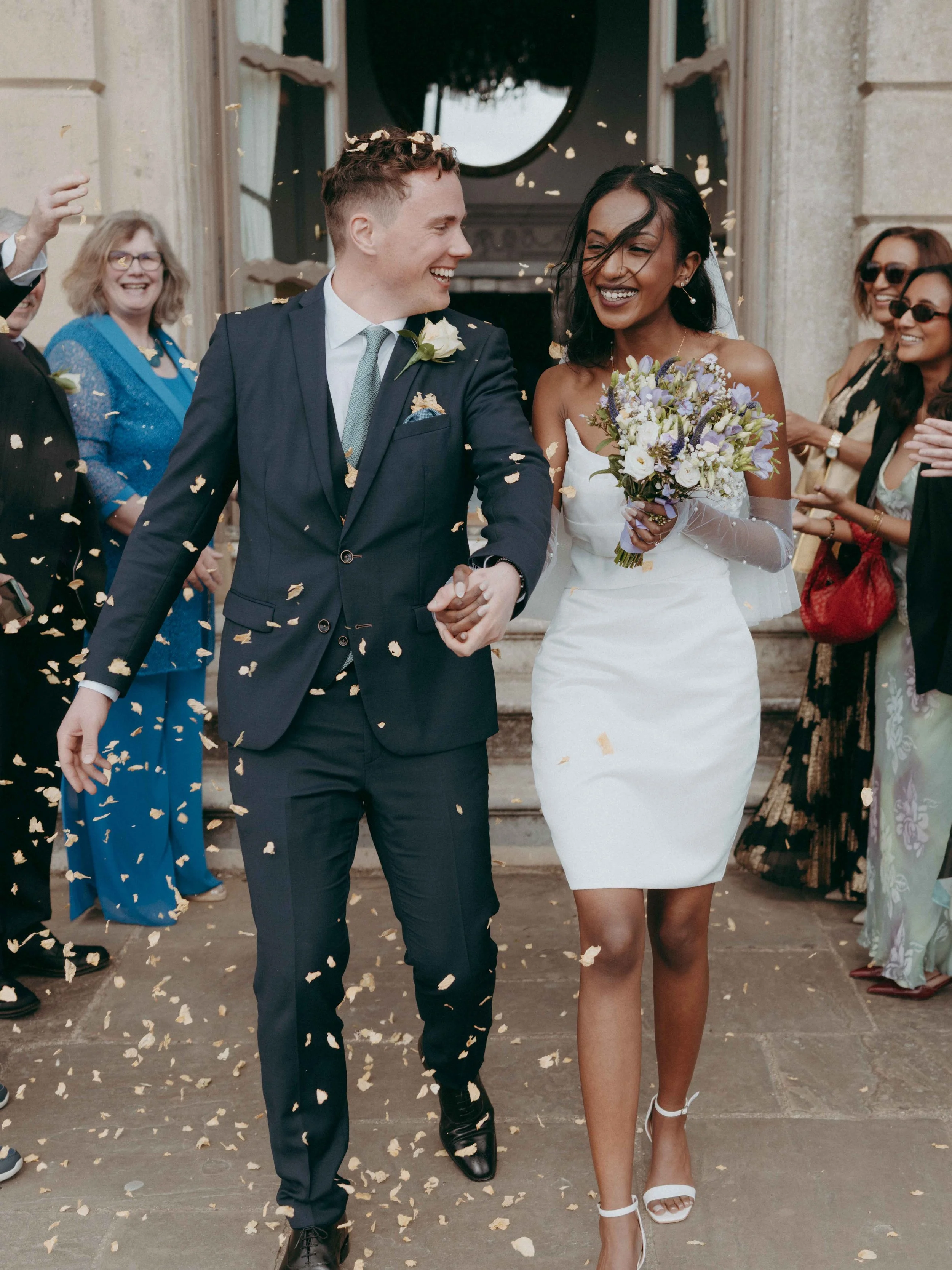 A newly married couple walks arm-in-arm outside a church, smiling, surrounded by wedding guests throwing confetti. Taken at Cliveden House wedding venue in Cliveden, Buckinghamshire, UK.