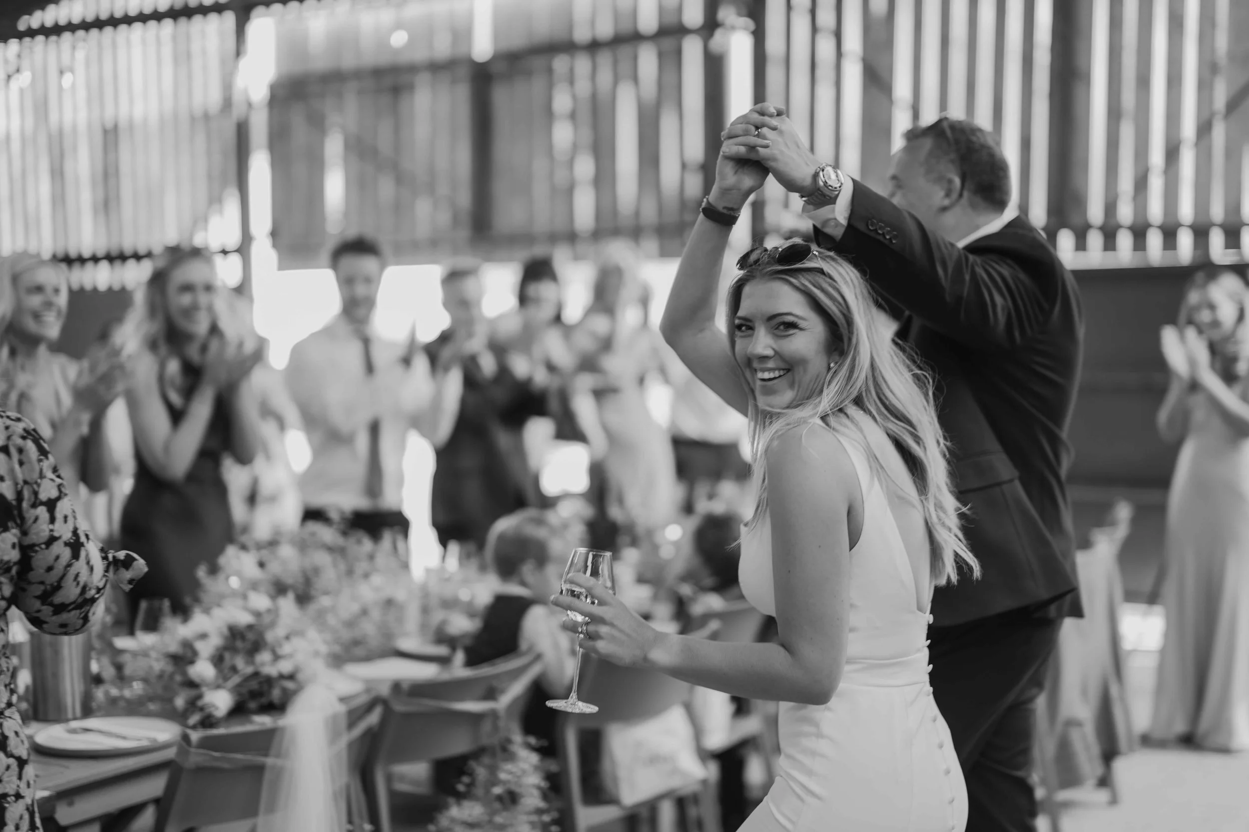 A newly wed couple's entrance to their reception, featuring a bride in a white dress smiling while holding a glass, and a groom in a black suit at a reception, surrounded by people clapping and celebrating. Taken at Artelium Wine Estate, Hassocks, UK